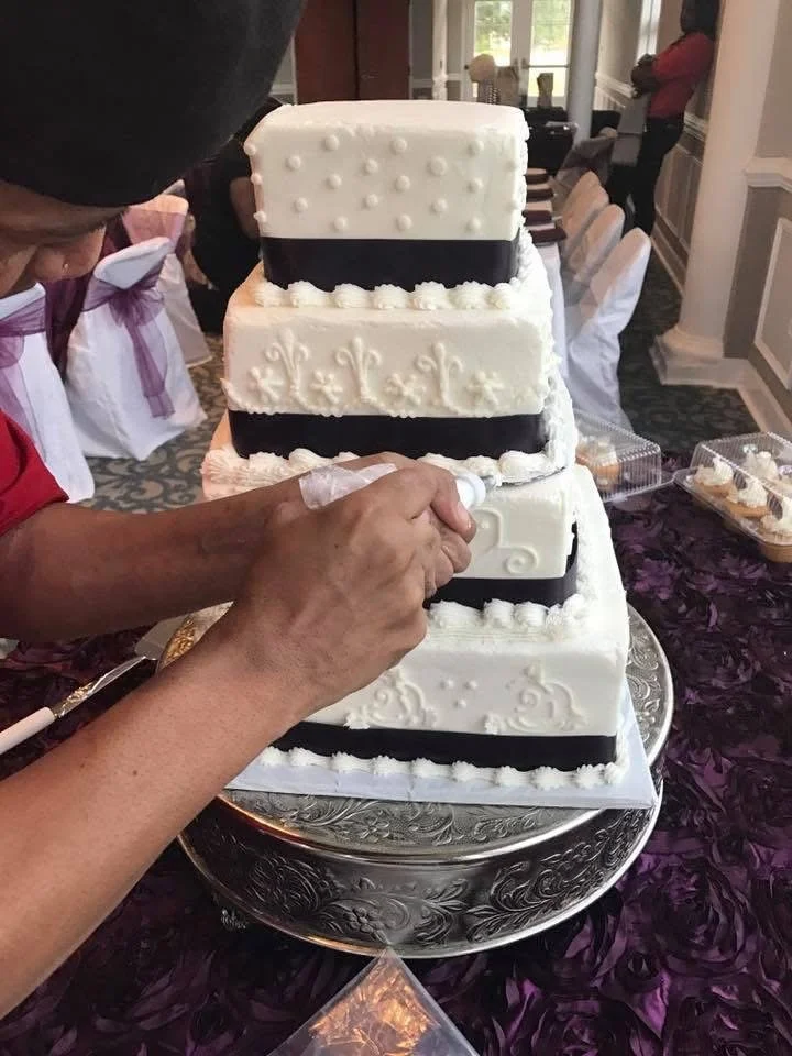 A person is cutting a white, four-tiered wedding cake with black ribbon accents and decorative icing patterns in a banquet hall.