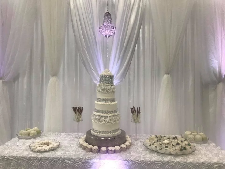 Elegant wedding cake with multiple tiers, decorative flowers, and embellishments on a decorated table with assorted desserts and props, backdrop with draped white fabric and a chandelier.