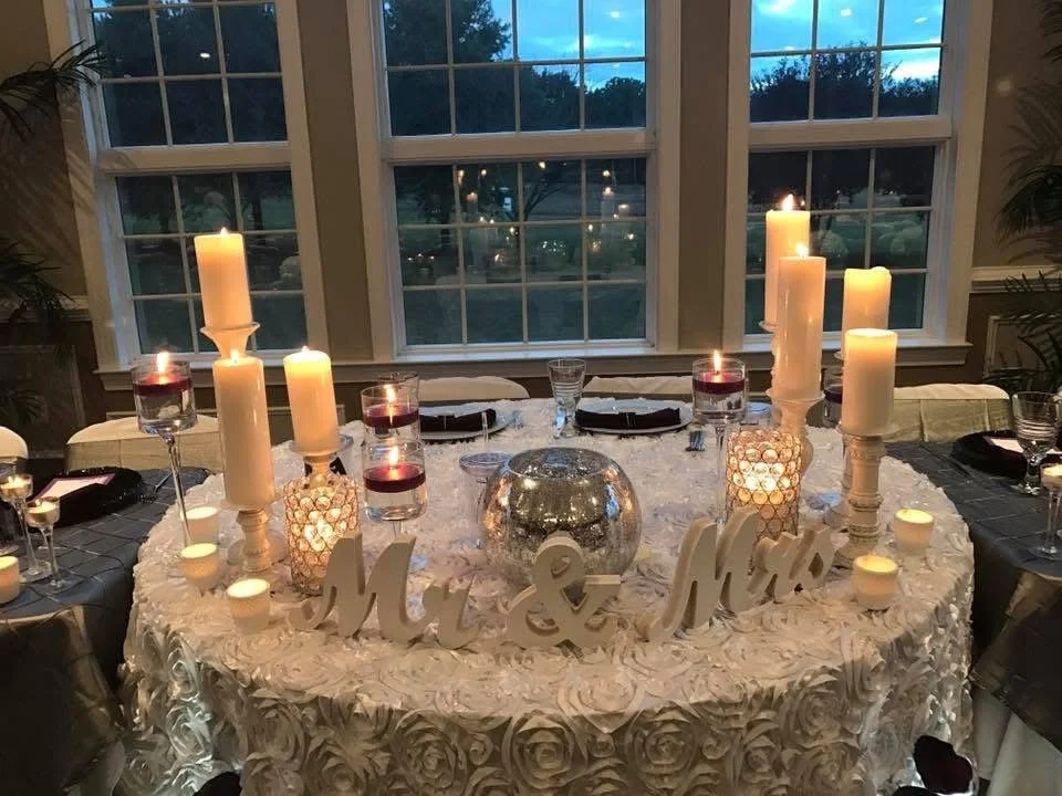 Wedding reception table decorated with white roses, large candles, smaller candles, and a glass centerpiece. The sign 'Mr. & Mrs.' is on the table, and the window reveals a view of trees outside during the evening.