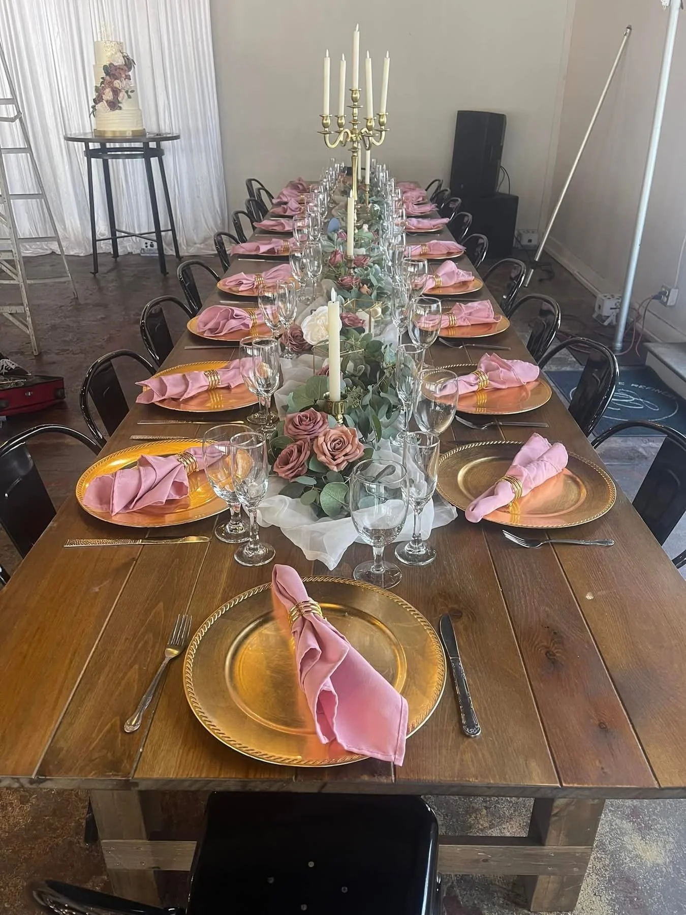 A long wooden dining table set for a formal event with pink cloth napkins, gold charger plates, wine glasses, silverware, a floral centerpiece with roses and greenery, and tall white candles in gold holders.