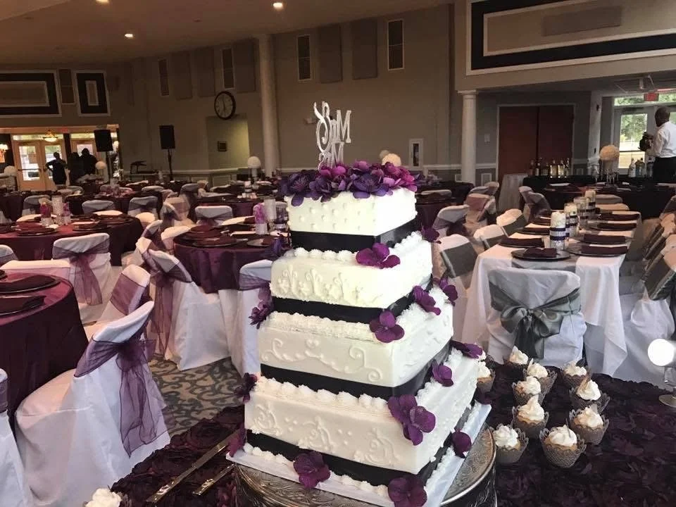Four-tier white wedding cake decorated with purple flowers and black ribbons, with a monogram cake topper, surrounded by cupcakes on a table in a banquet hall.