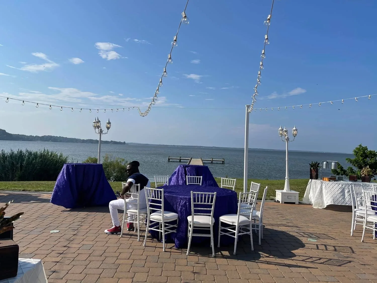 Outdoor event setup by a waterfront with tables draped in purple and white cloths, white chairs, string lights, and decorative lamp posts under a blue sky with scattered clouds.