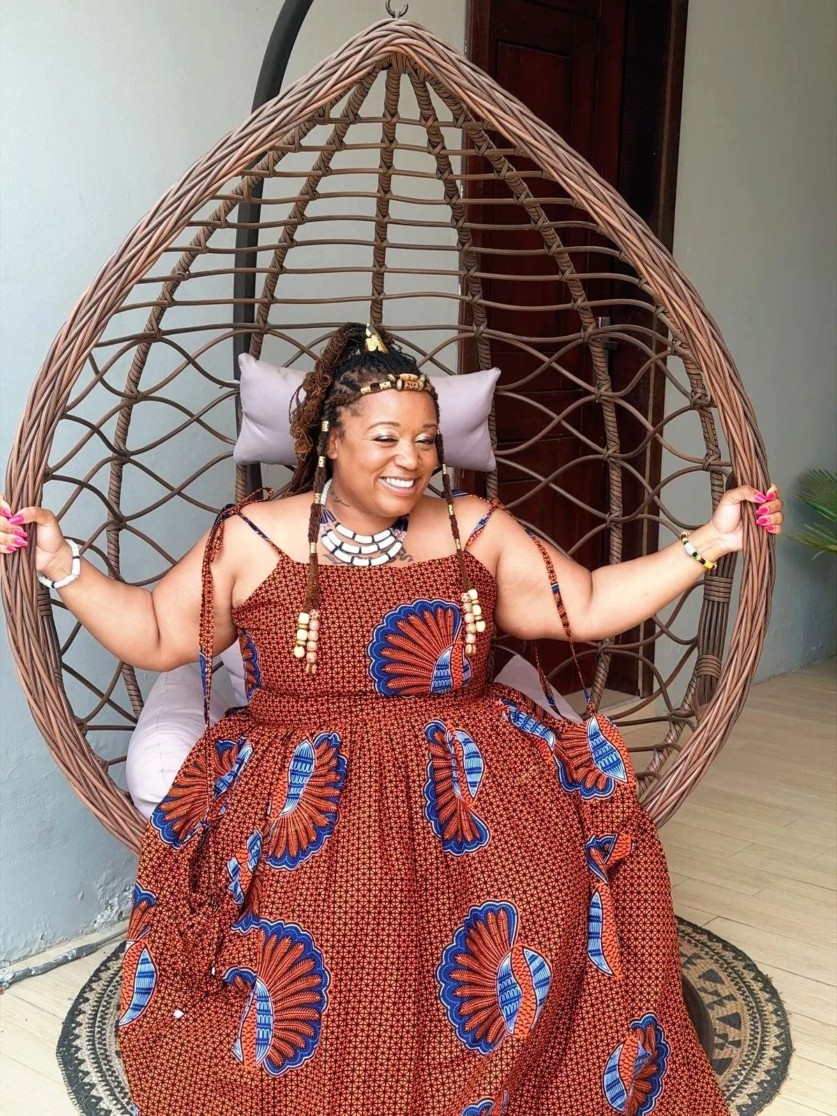 Woman sitting in a round, wicker hanging chair with a gray pillow, smiling, wearing a colorful, patterned dress and accessories, indoors with a wooden door and a plant in the background.