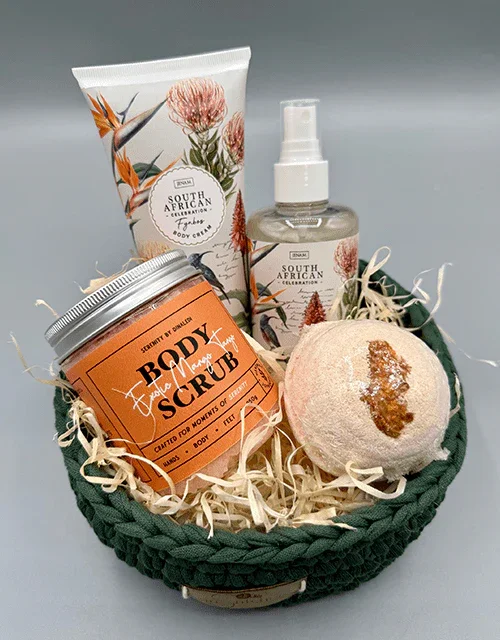 A gift basket containing a body scrub jar, a spray bottle, a tube with floral design, and a bath bomb, all arranged on shredded paper in a dark green woven basket.