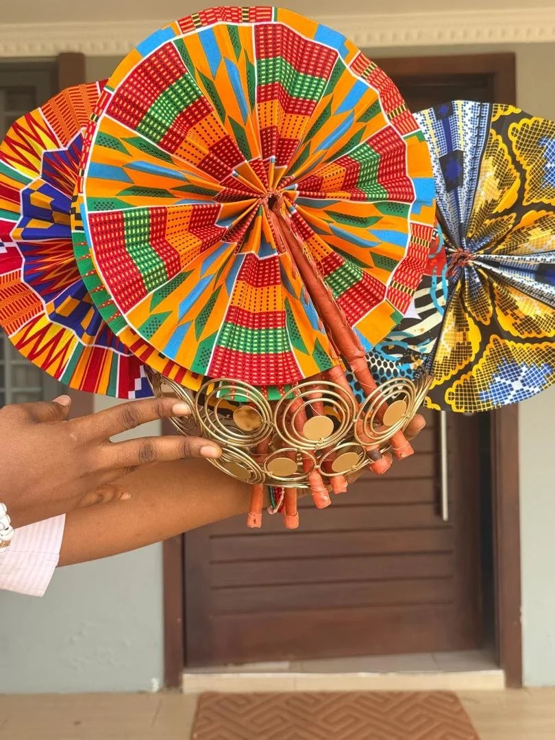 Multiple colorful traditional African umbrellas with intricate patterns are held together by a person. The umbrellas feature vibrant colors such as orange, red, green, yellow, and blue, with decorative gold elements at the base.