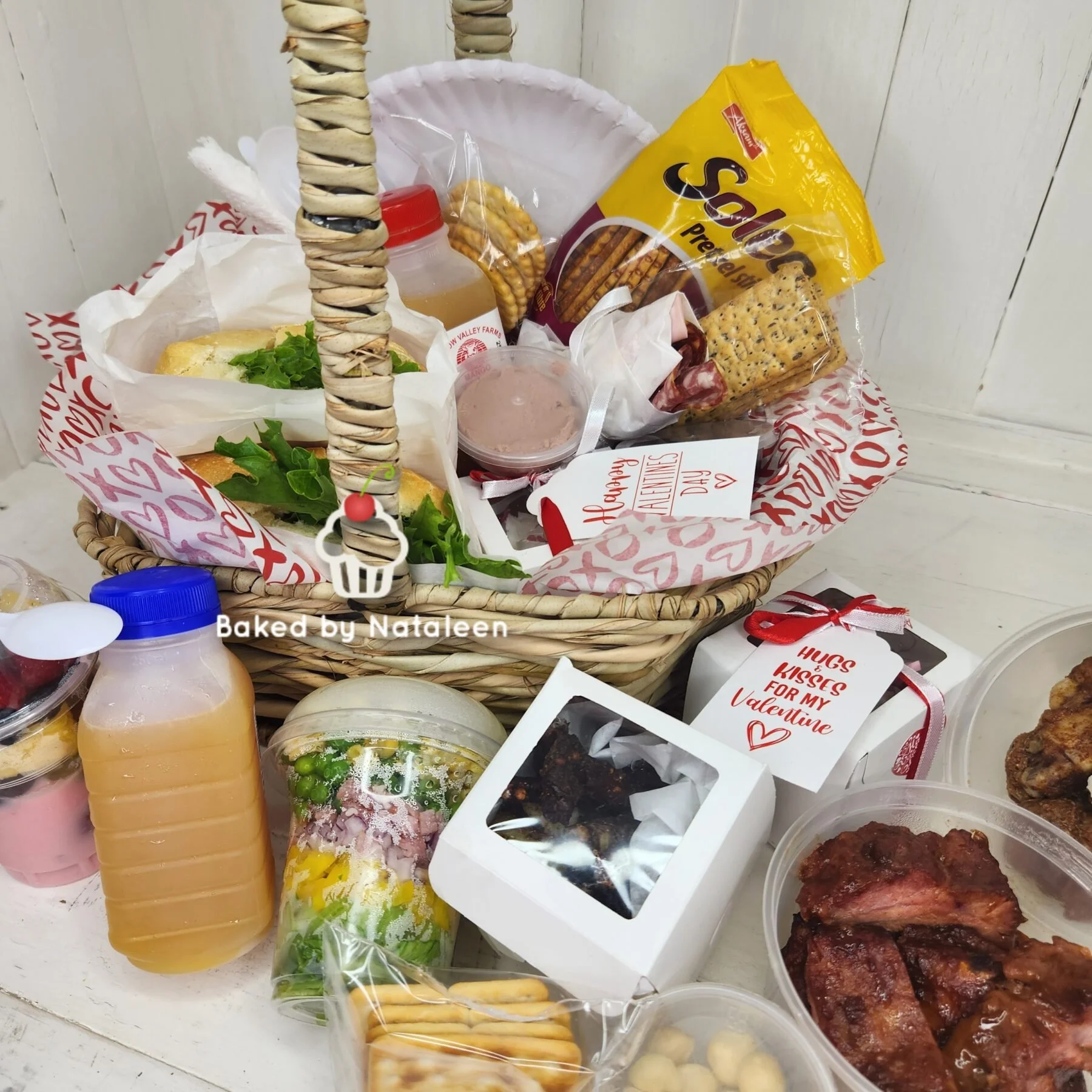 A Valentine's Day gift basket filled with snacks, sandwiches, cookies, drinks, and chocolates, accompanied by a small container of chicken wings and a decorated box of chocolates.