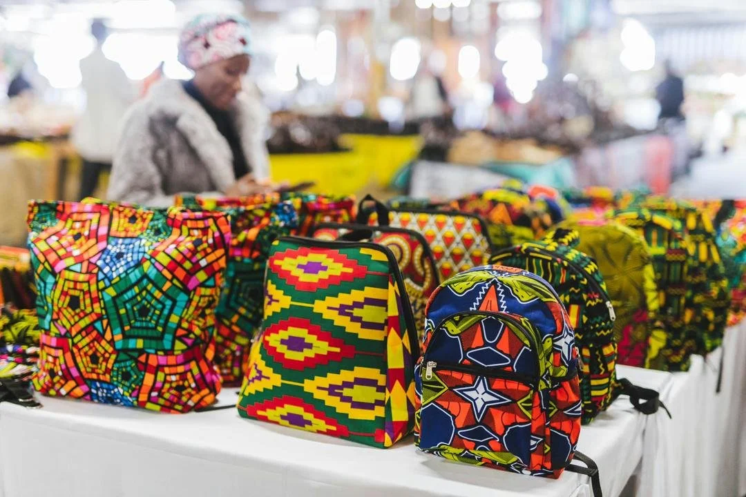 Colorful patterned bags and backpacks displayed on a white table at a market or fair. A woman wearing a headscarf and fur coat is browsing in the background.
