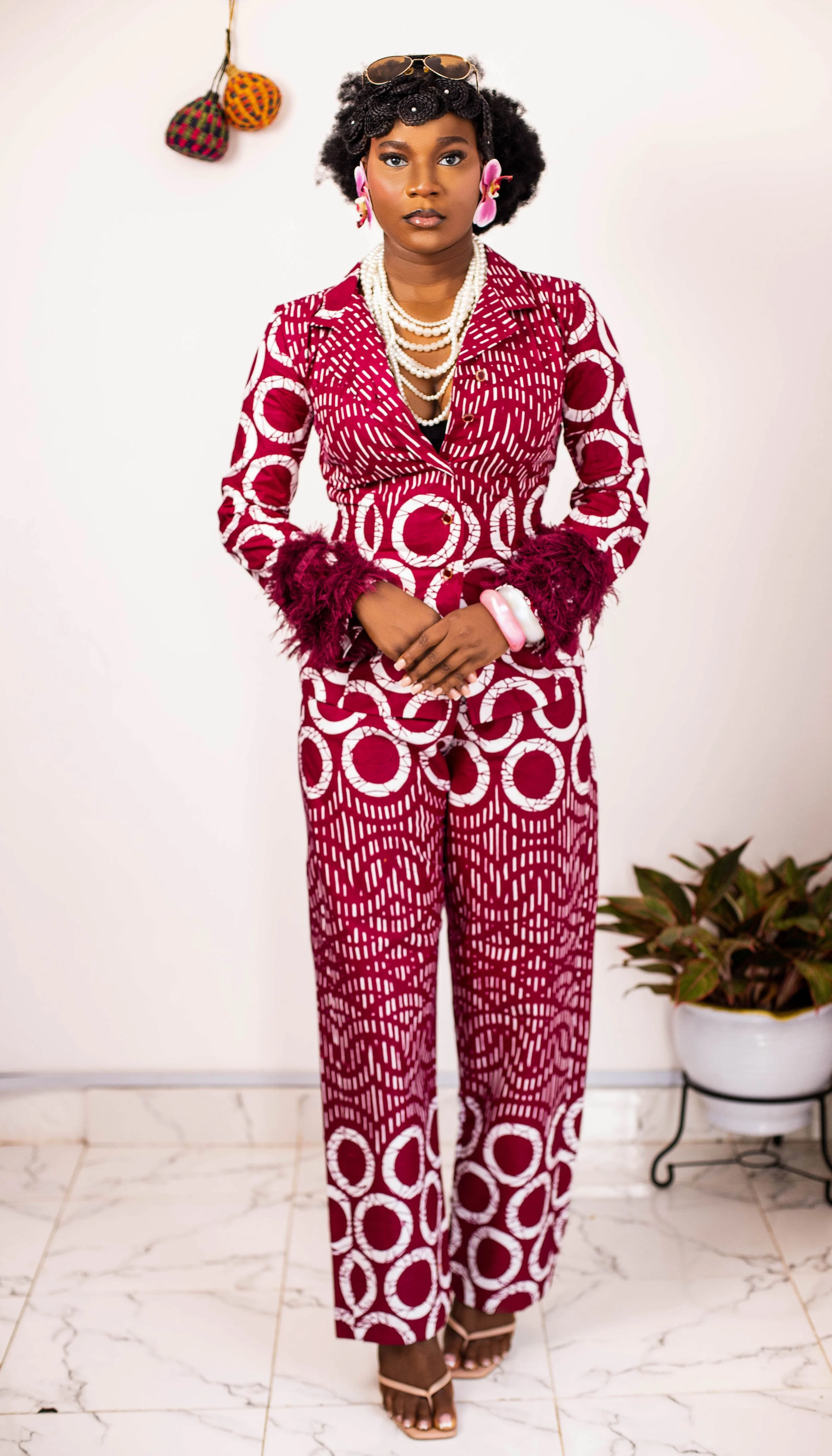 A woman wearing a red and white patterned suit with purple accents, standing in front of a plain white wall. She has curly black hair, pink floral earrings, layered pearl necklaces, and a pair of sunglasses on her head. She is accessorized with chunky bracelets and wears open-toed heels. There is a potted plant on the right and a decorative wall hanging on the left.