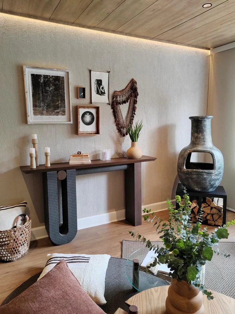 Living room corner with decorated wall, tall ceramic vase on a stand, and a wooden table with a vase of greenery.