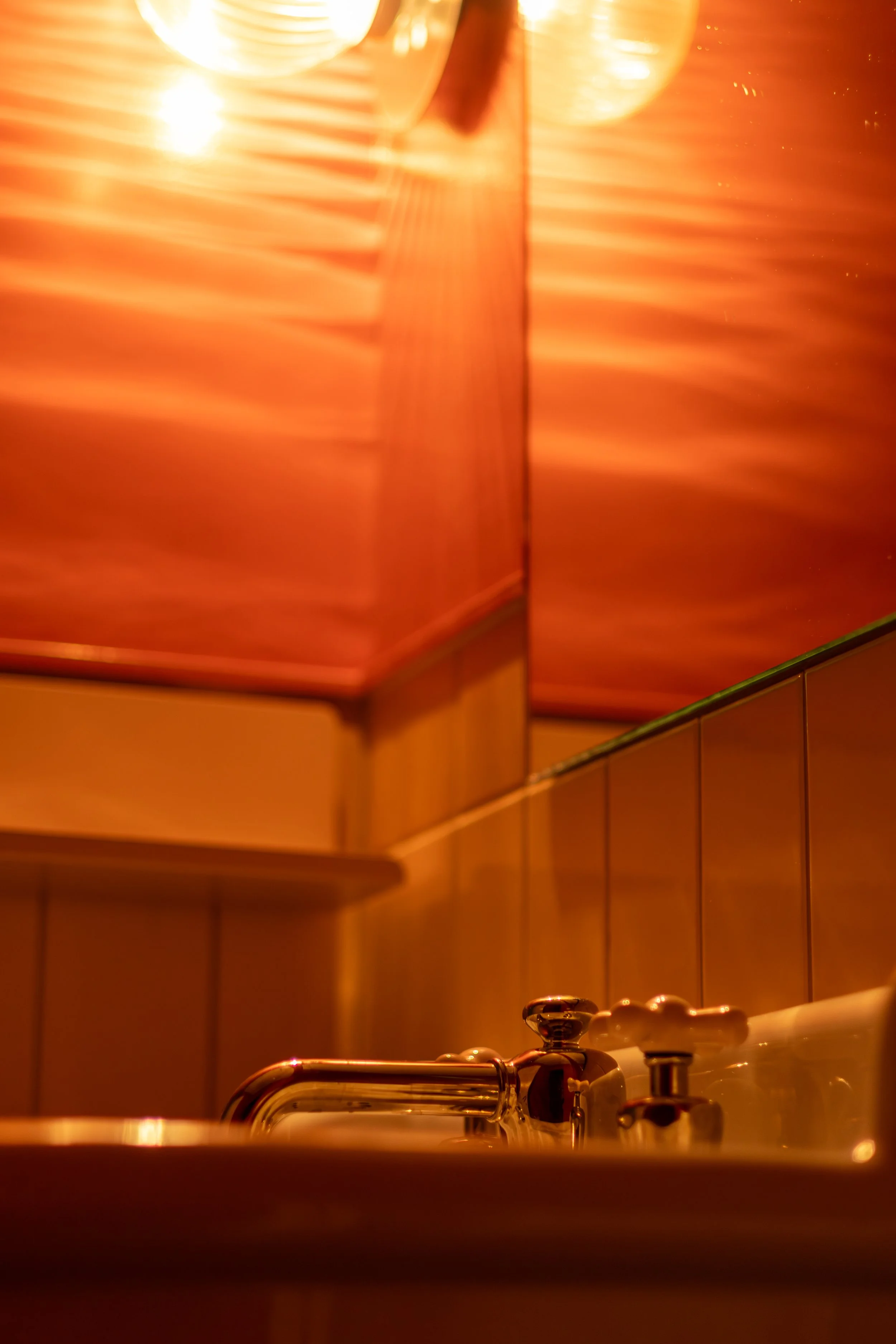 Close-up of a bathroom sink with a chrome faucet and soap dispenser, with warm lighting and orange background reflections.