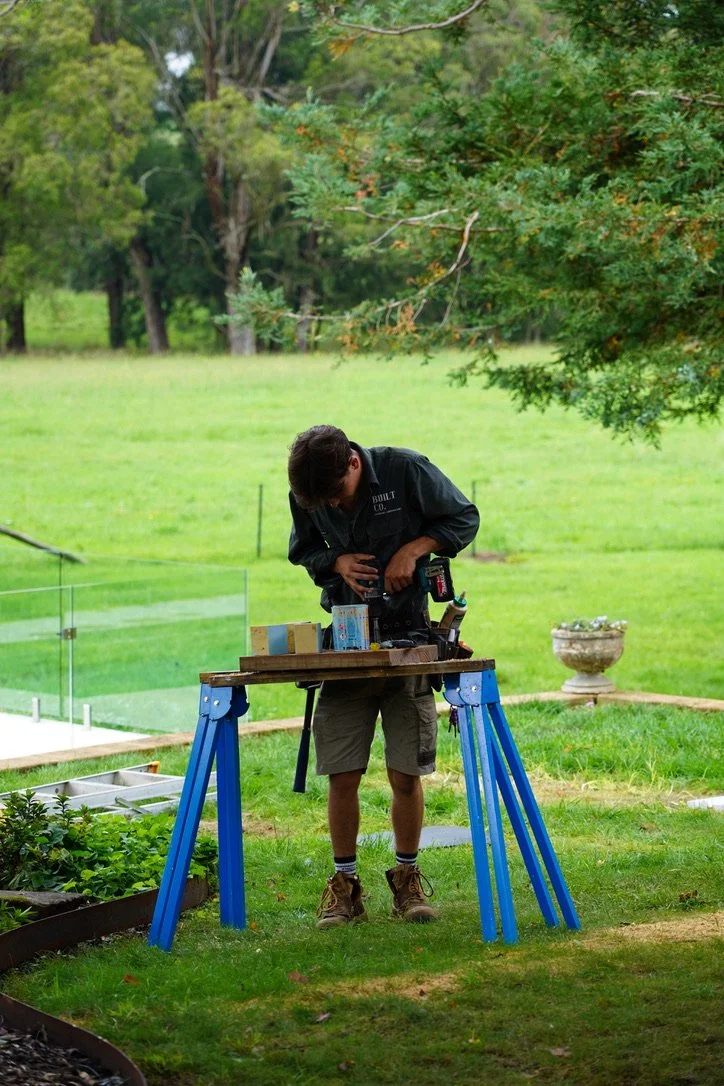 A man working on a DIY project outdoors in a green backyard, using a drill on a wooden workbench.