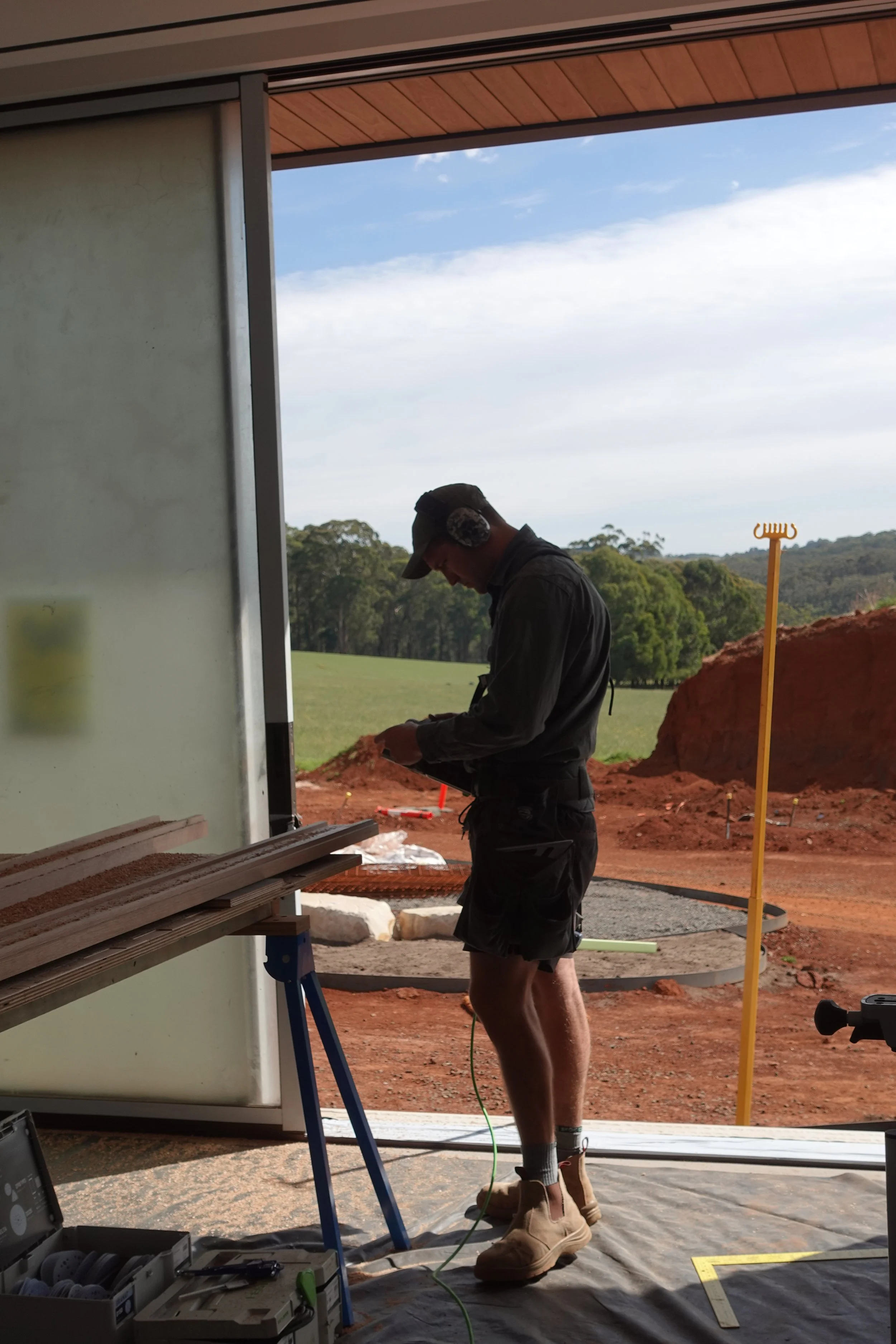A man wearing headphones, a cap, and work boots, standing inside near an open door at a construction site, looking at a mobile device. Outside are trees, dirt, and construction materials.