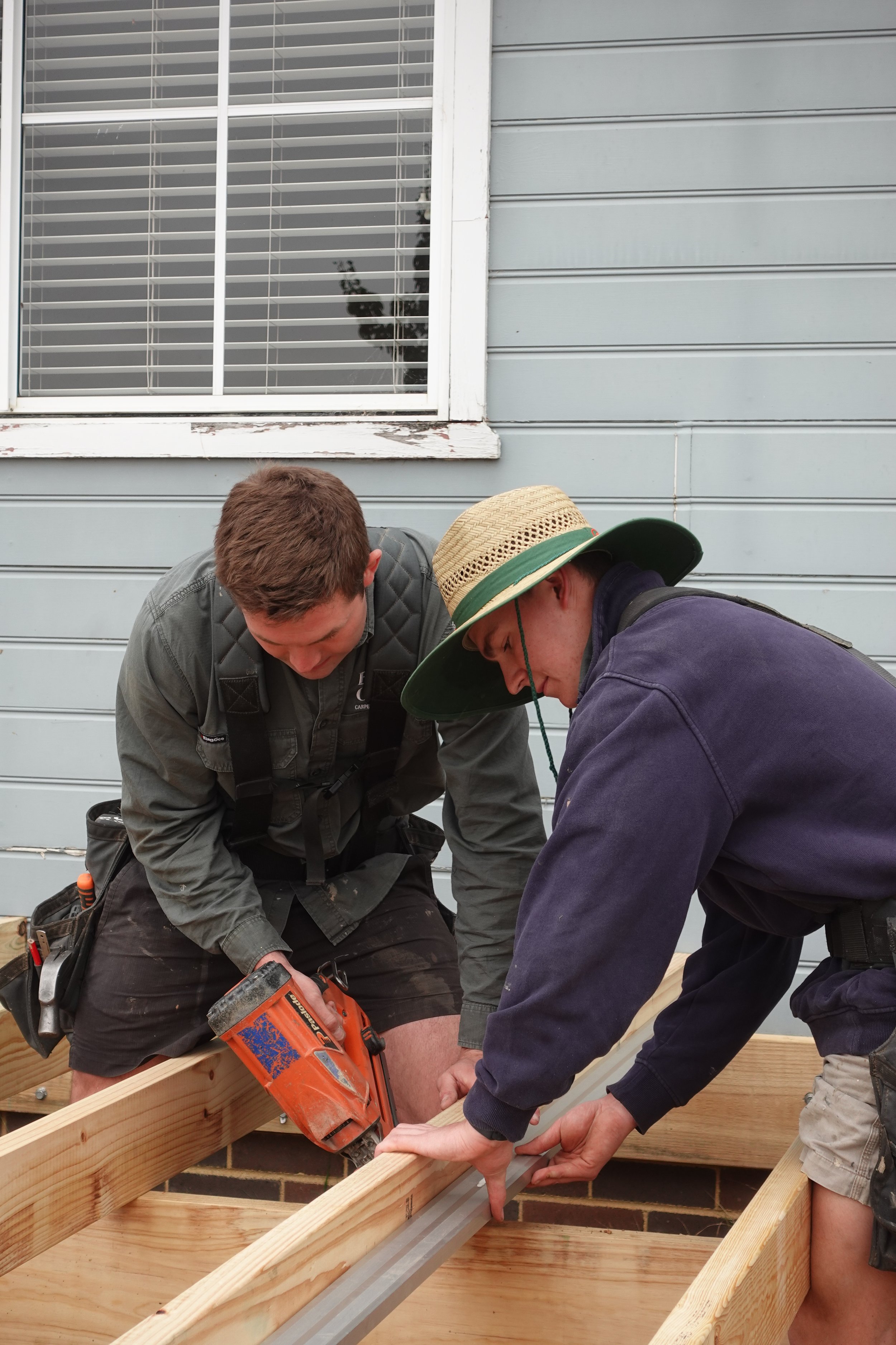 Two men working together on a construction project, measuring and aligning a piece of lumber on a wooden frame outside a house.