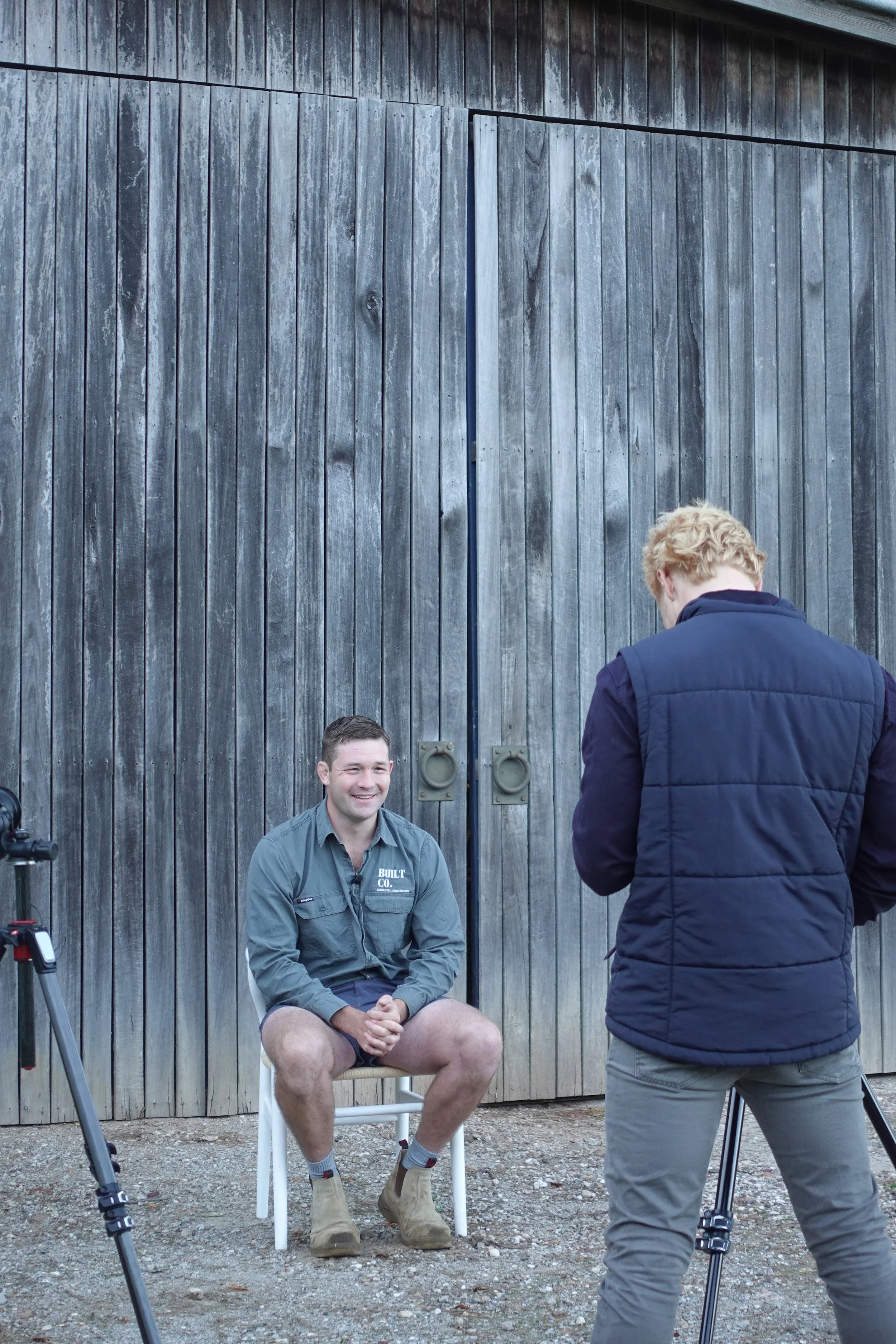 A man sitting on a white chair in front of a large wooden door, smiling during a photoshoot or interview. A tripod is visible on the left, and another person stands with their back to the camera, facing him.