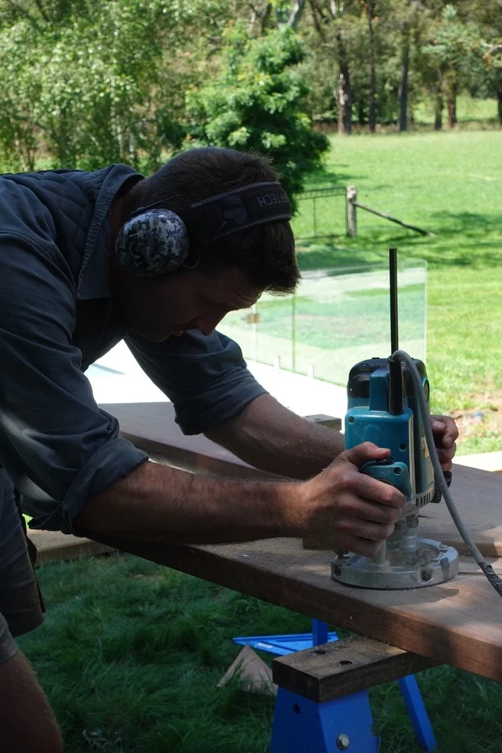 A man wearing headphones and a dark shirt is using a power router on a wooden board outdoors, with grass, trees, and a fence in the background.