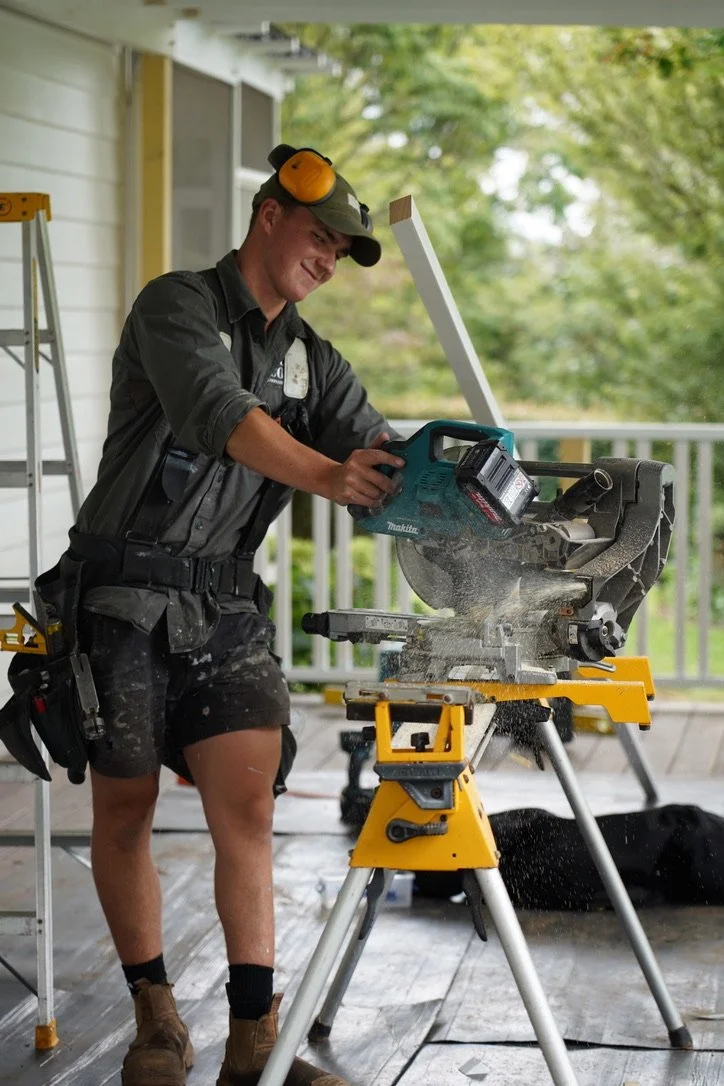 A man working with a miter saw on a porch, wearing safety gear including headphones and a cap, with trees in the background.