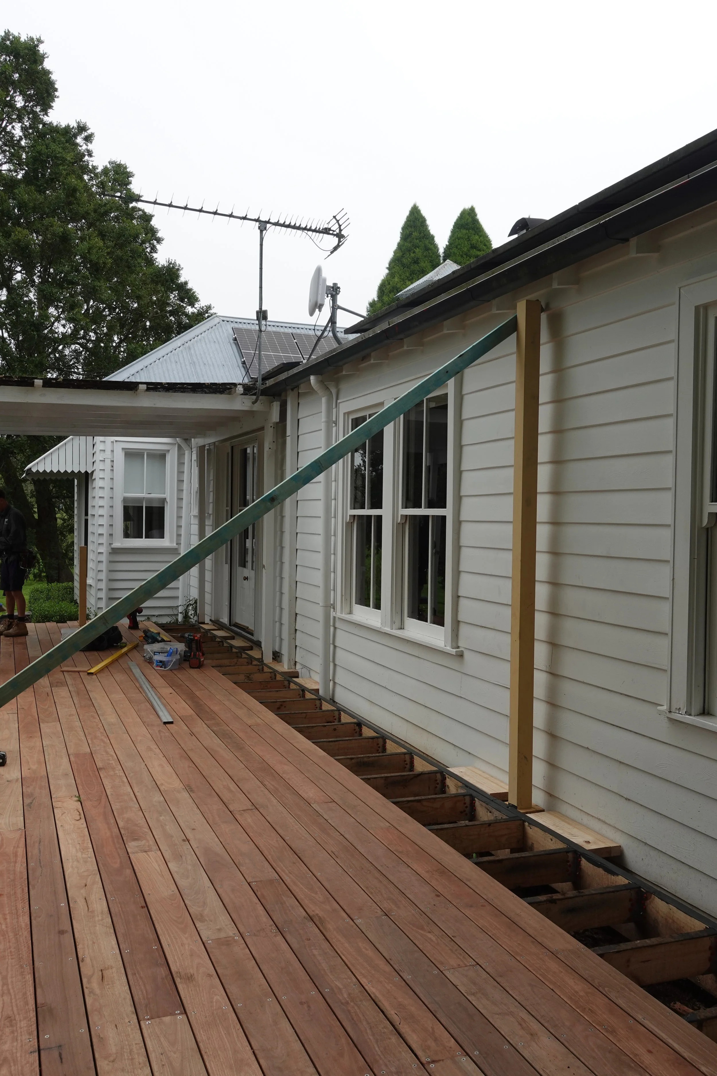 Construction of a wooden deck attached to a house, with tools and materials scattered on the deck, and support posts in place for railing installation.