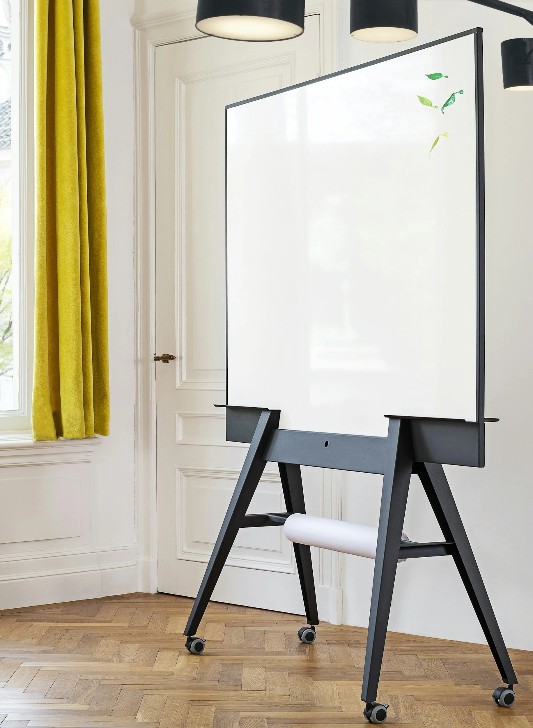 A modern whiteboard on a black stand with wheels in a room with wooden flooring, a window with yellow curtains, and black and white lighting fixtures.