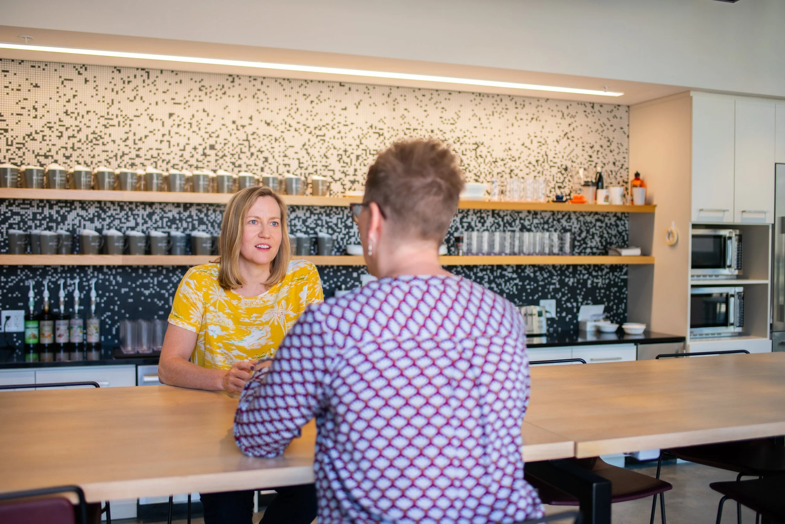 A woman with shoulder-length blonde hair and a yellow floral dress sitting at a counter talking to a person with short hair wearing glasses and a patterned shirt in a modern cafe with black and white patterned wall and shelves with cups and bottles.