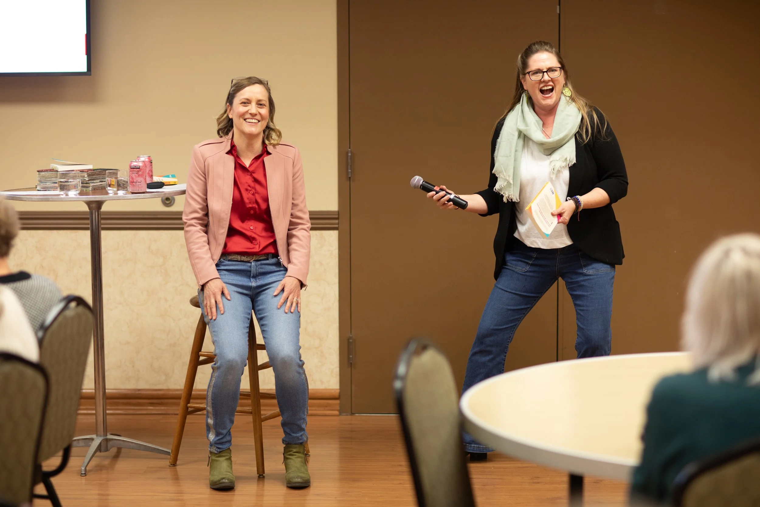 Two women at a presentation or workshop, one sitting on a chair and smiling, the other standing and speaking into a microphone with a joyful expression.