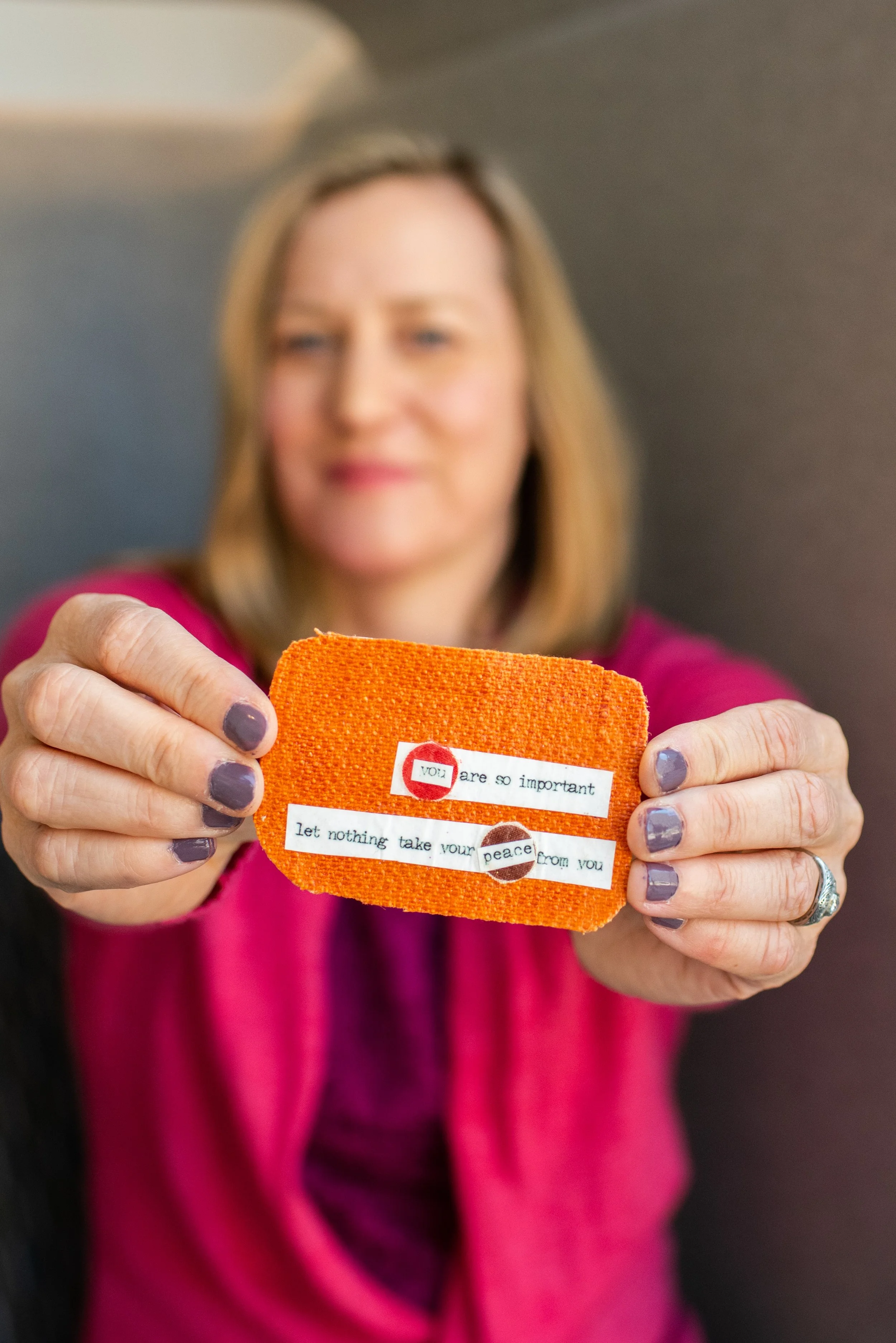 A woman with shoulder-length blonde hair holding an orange heart-shaped card with a message, wearing a pink top, with a blurred background.