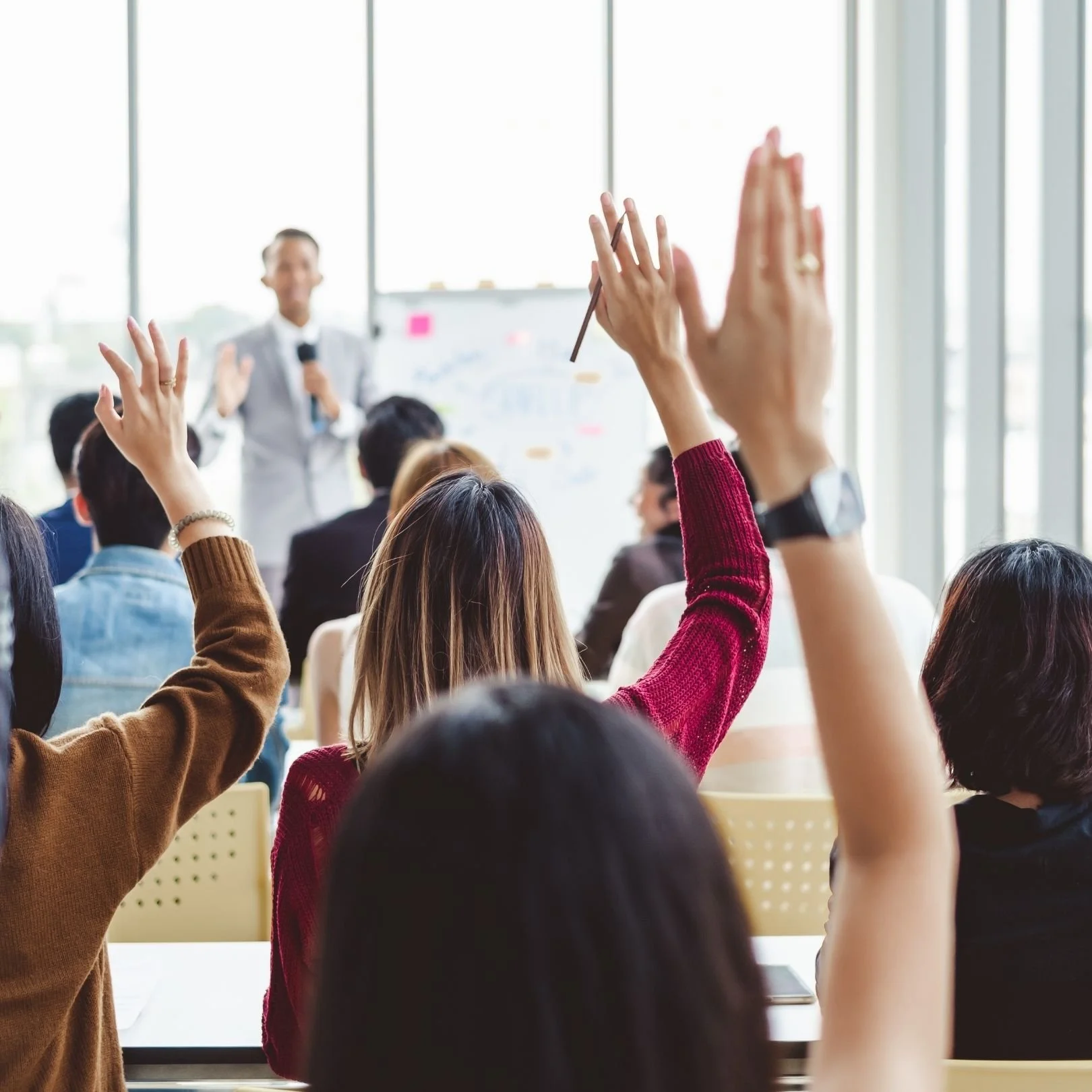 Students in a classroom raising their hands, with a blurred teacher at the front near a whiteboard.