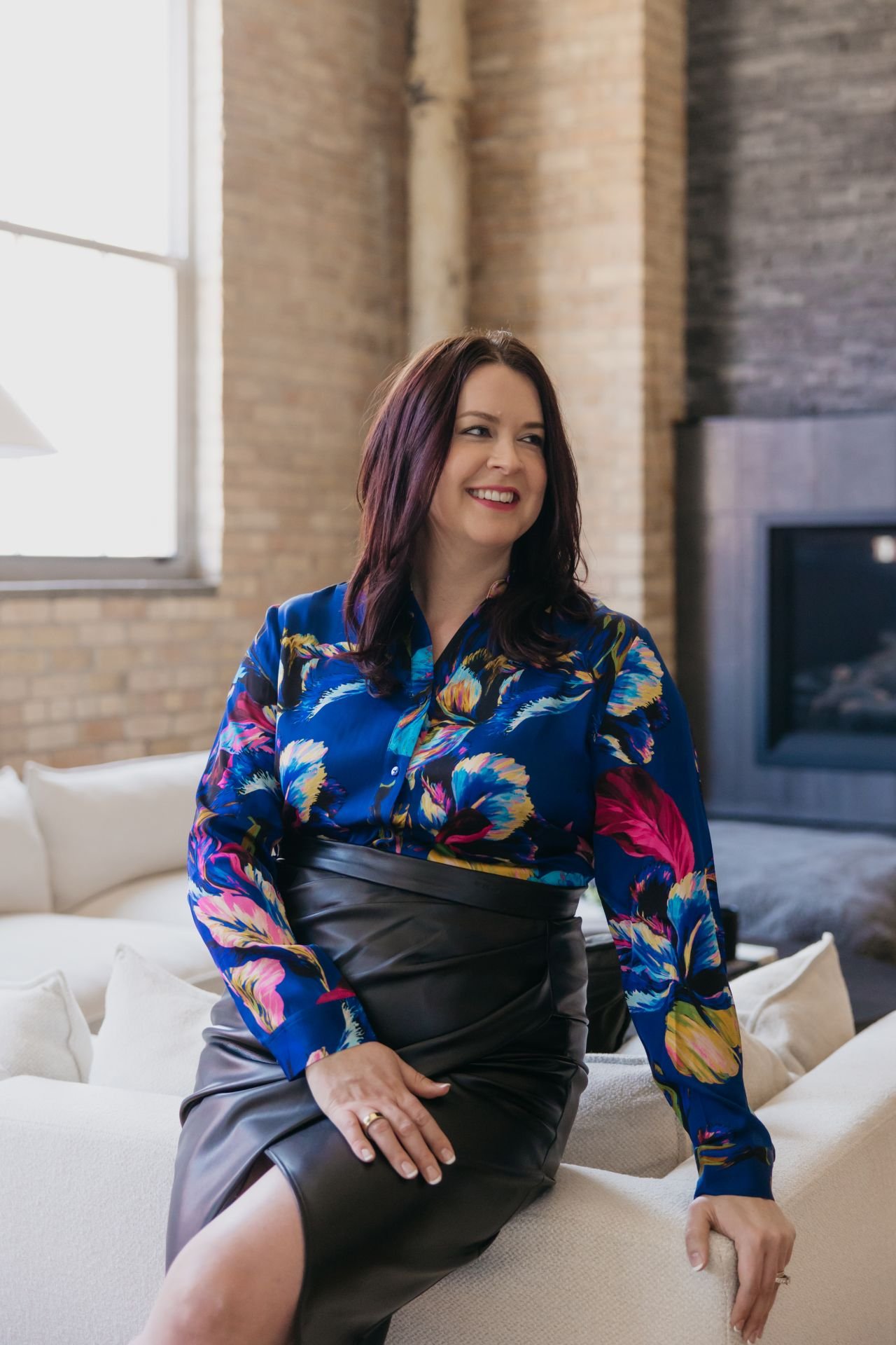A woman with long dark hair smiling and sitting on a white sofa in a modern, industrial-style living room with brick walls and large windows.