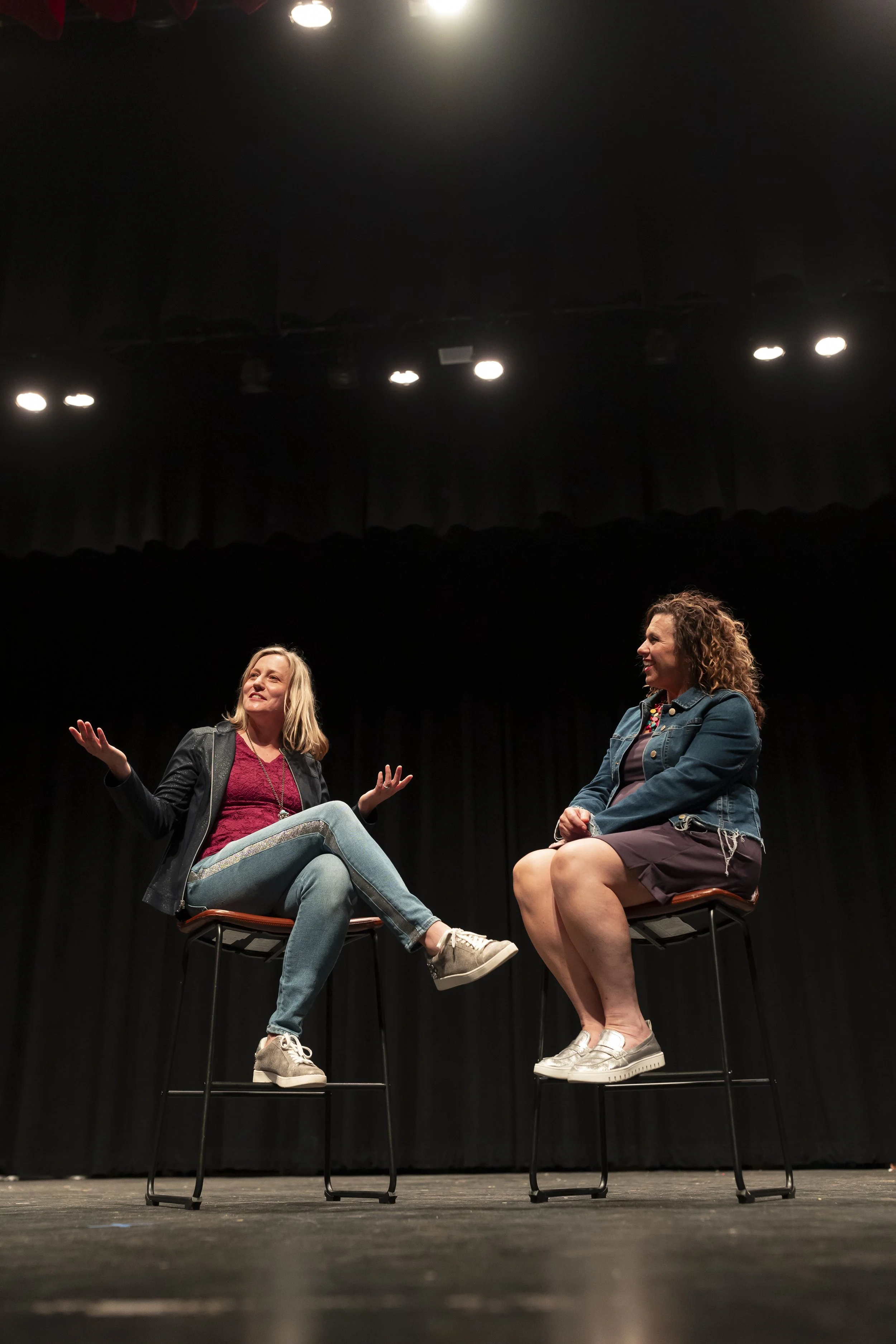 Two women are sitting on chairs on a stage, engaging in a conversation. The woman on the left has blonde hair, is gesturing with her hands, and is wearing a dark jacket, red top, and light jeans. The woman on the right has curly hair, is smiling, and is wearing a denim jacket and a dress. There are stage lights and a black curtain in the background.