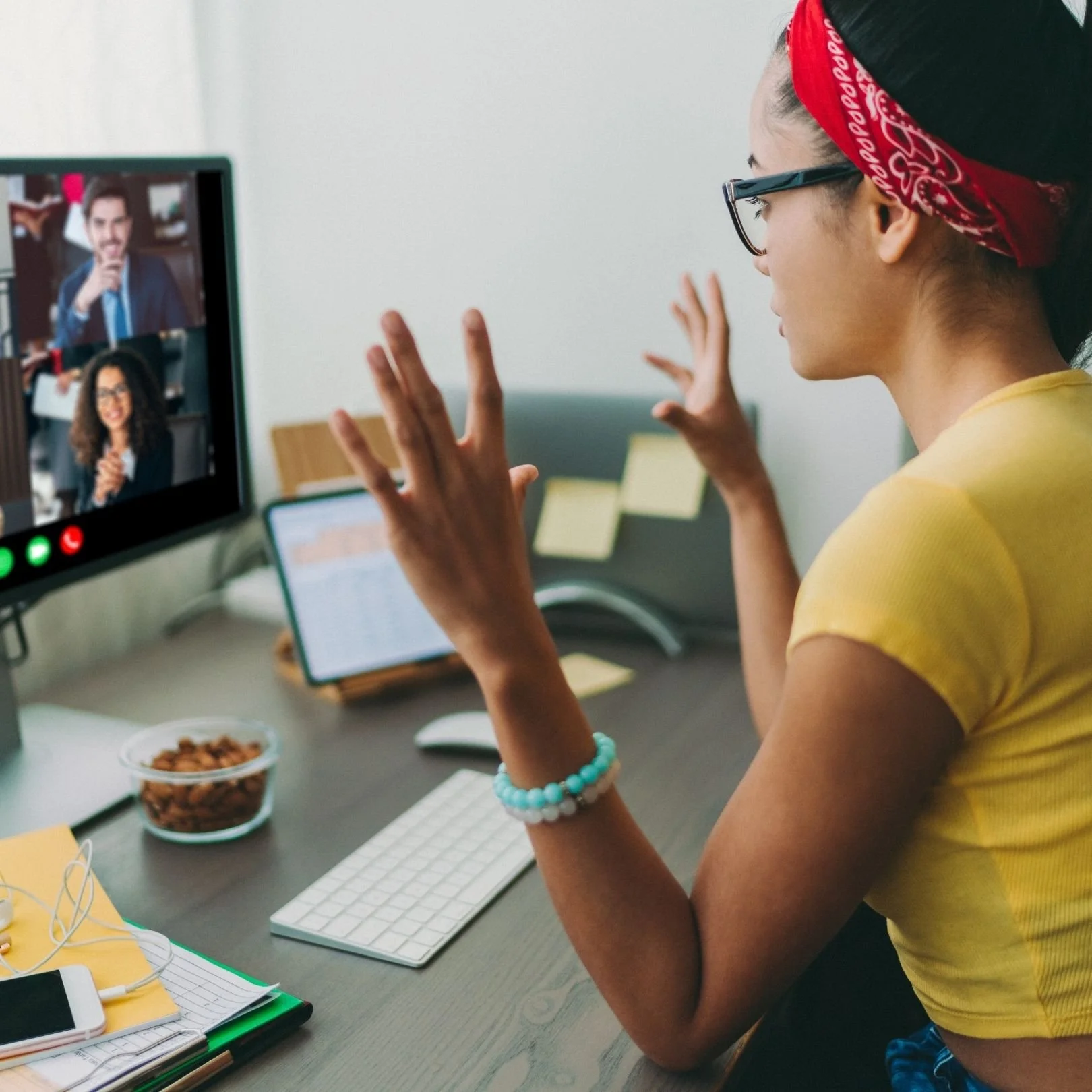 A young woman with glasses and a red bandana is waving during a video call on her computer. She is sitting at a desk with a keyboard, a bowl of snacks, notebooks, and a smartphone.