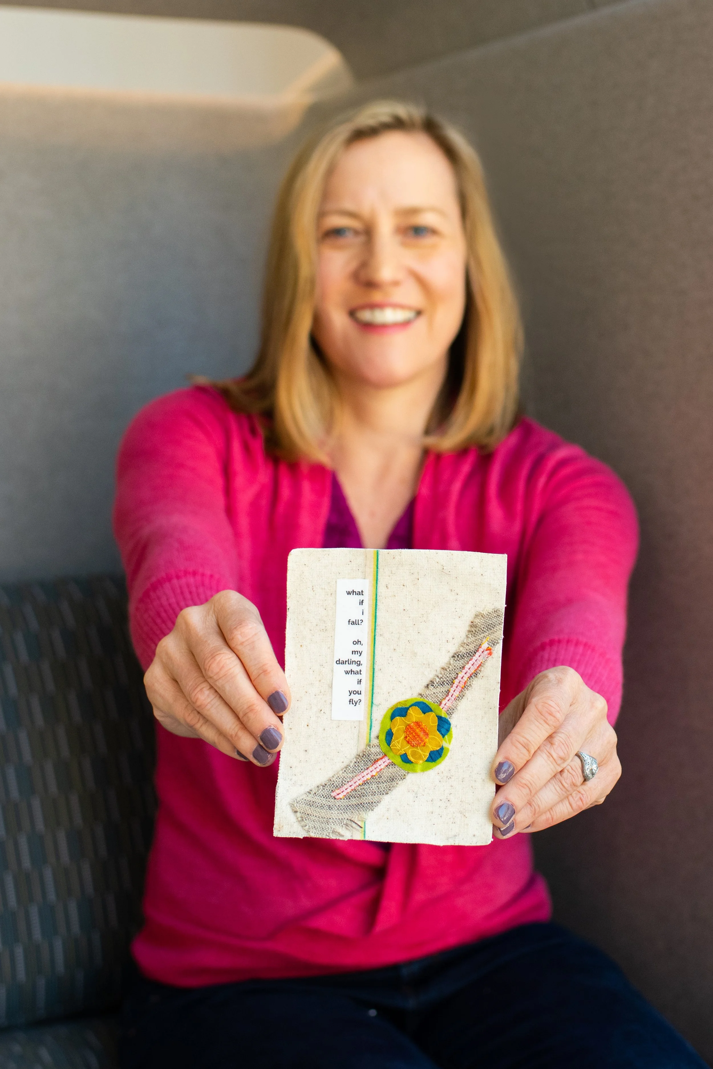 A woman in a pink sweater holds a handmade fabric card decorated with a felt flower, ribbon, and features a poem. She is seated on a patterned chair.