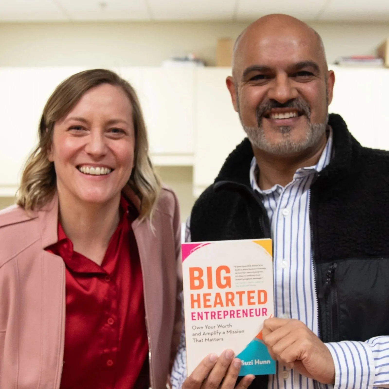 Smiling woman in a pink jacket and a smiling man holding a book titled 'Big Hearted Entrepreneur' at an indoor event.