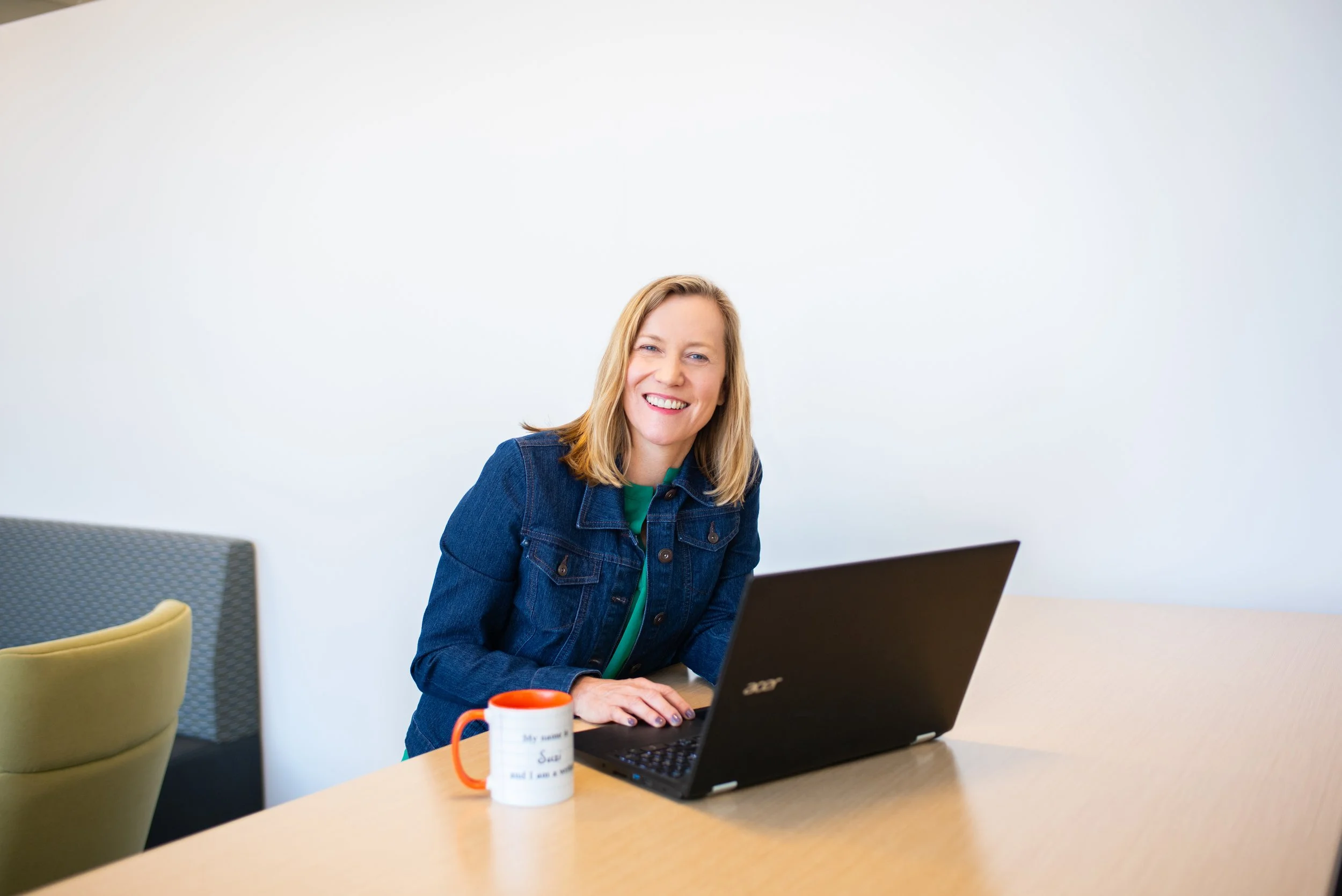 A woman with blonde hair, wearing a denim jacket, sitting at a table with a laptop and a white mug with orange handle, smiling at the camera against a plain white wall.