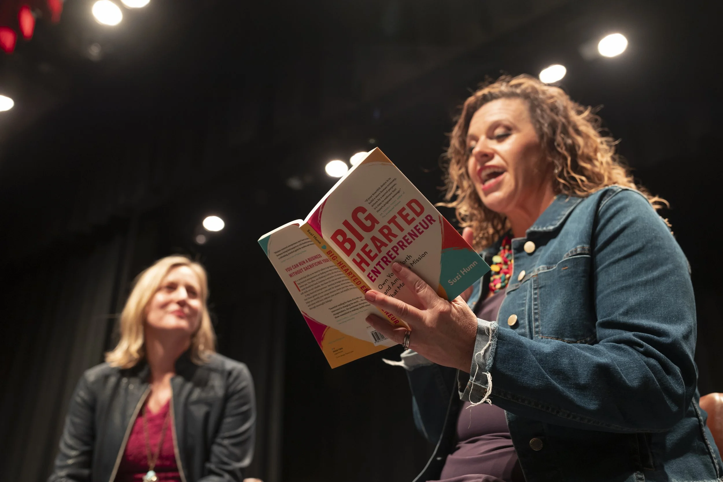 A woman with curly hair, wearing a denim jacket and colorful necklace, is reading a book titled "Big Hearted Entrepreneur" at an indoor event. Two women are visible in the background, one with straight blonde hair and the other with wavy hair, in a dimly lit setting with overhead lights.