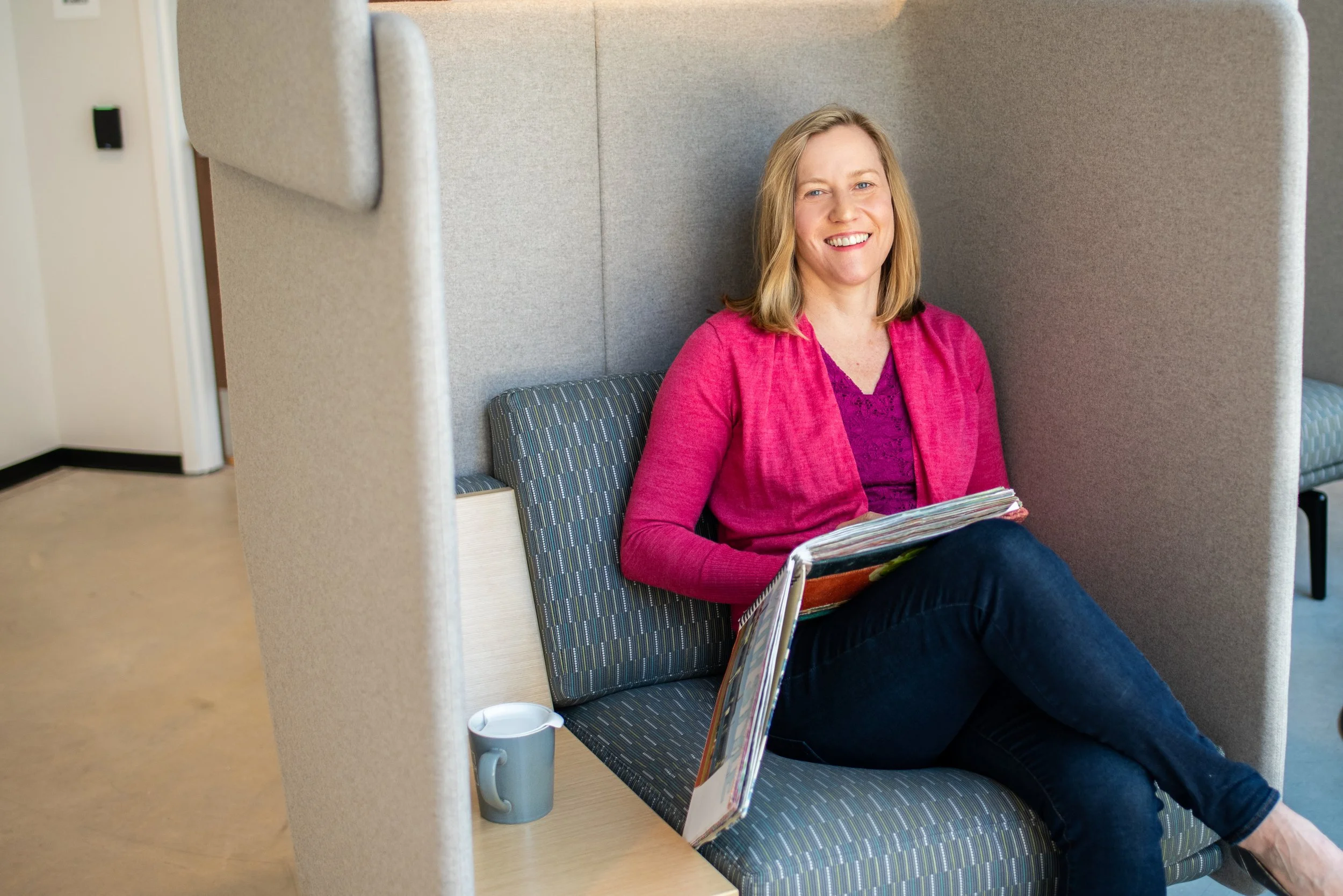 A woman with blonde hair smiling, sitting in a private booth at a public space, wearing a pink blazer over a purple top, holding a magazine. There is a white mug on the table in front of her.