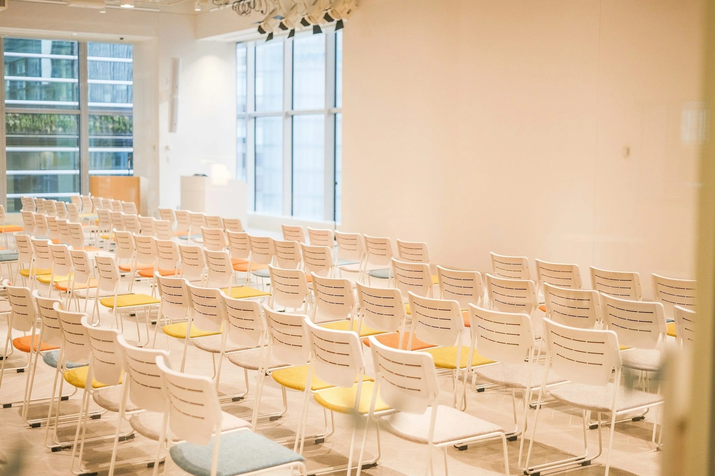 Empty chairs arranged in rows in a modern, well-lit conference room with large windows and beige walls.