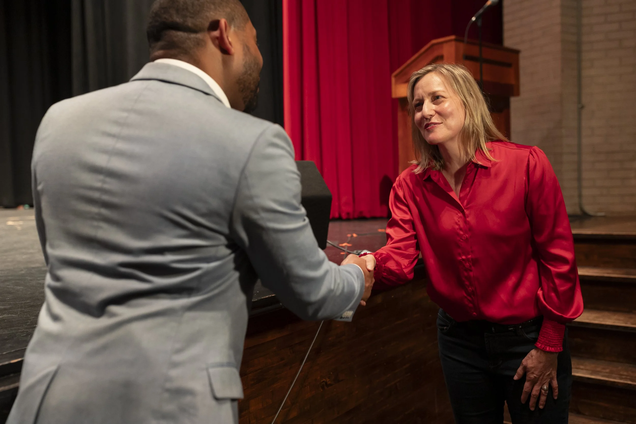 A man in a light gray suit shaking hands with a woman in a red blouse on a stage with red curtains in the background.
