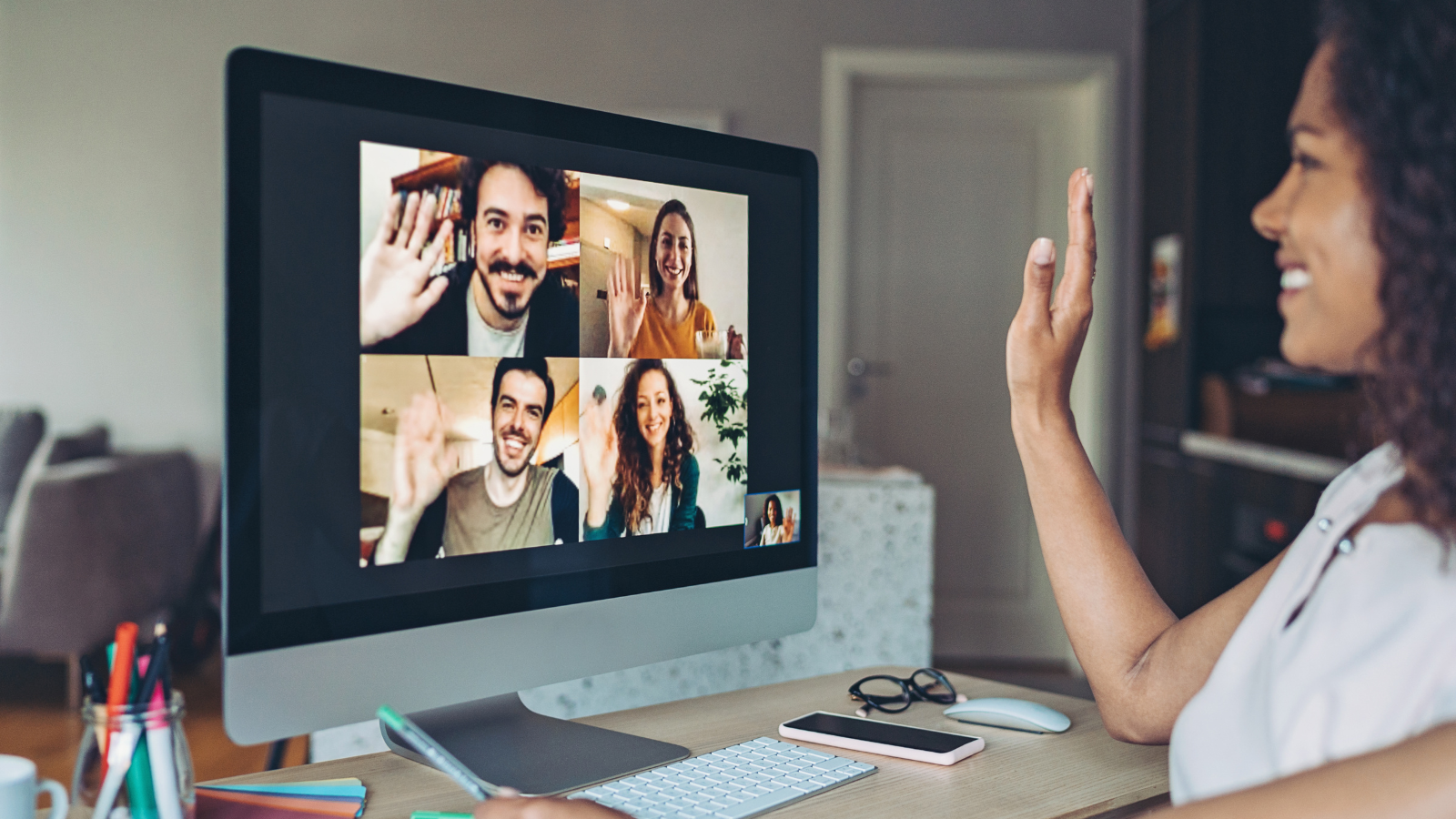 A woman participating in a video call with five other people, all waving and smiling on the computer screen.