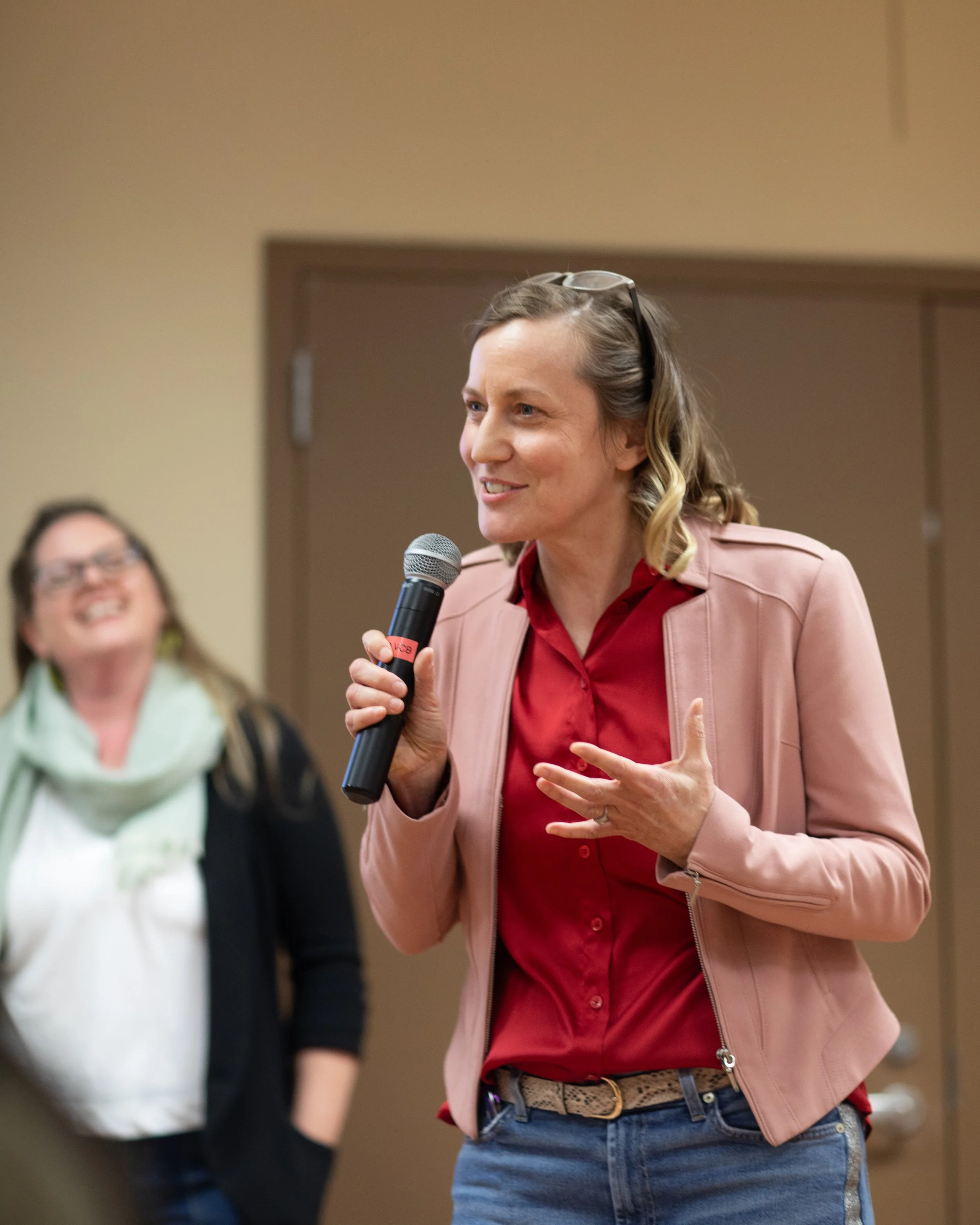 Woman with glasses on her head speaking into a microphone in a room with beige walls and a wooden door, while a woman in glasses and a scarf listens in the background.