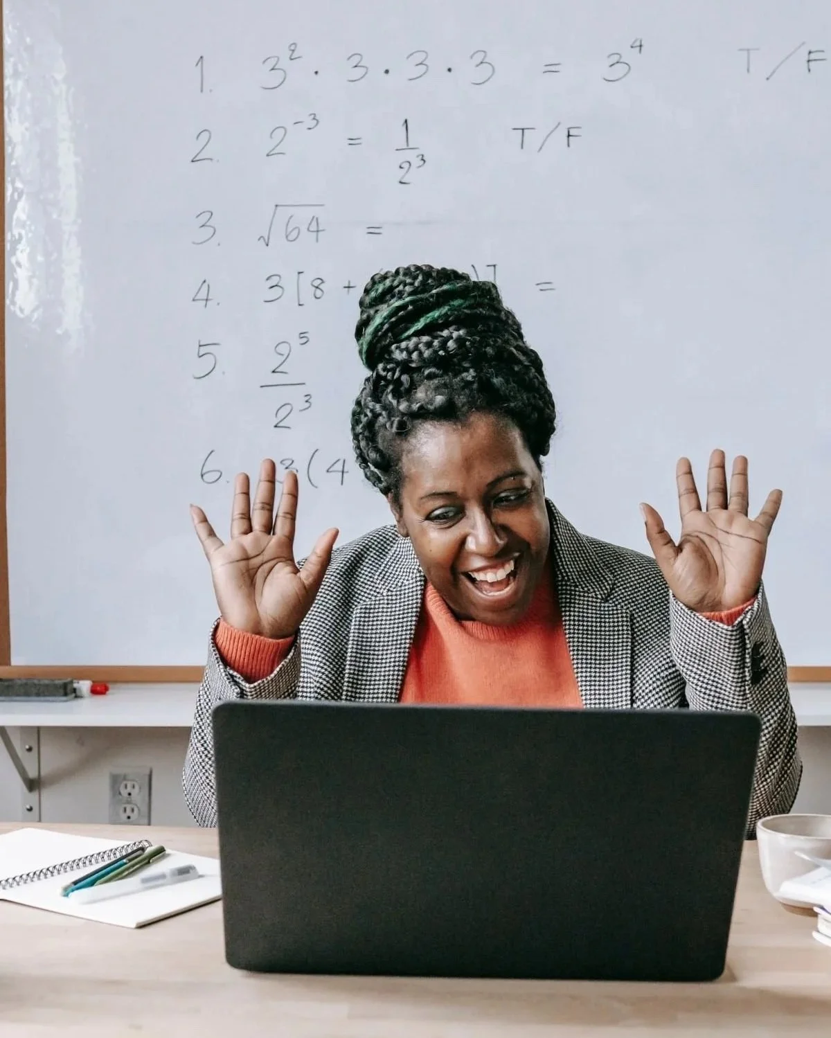 A woman smiling and playing with her hair in front of a laptop, with a whiteboard filled with handwritten math problems in the background.