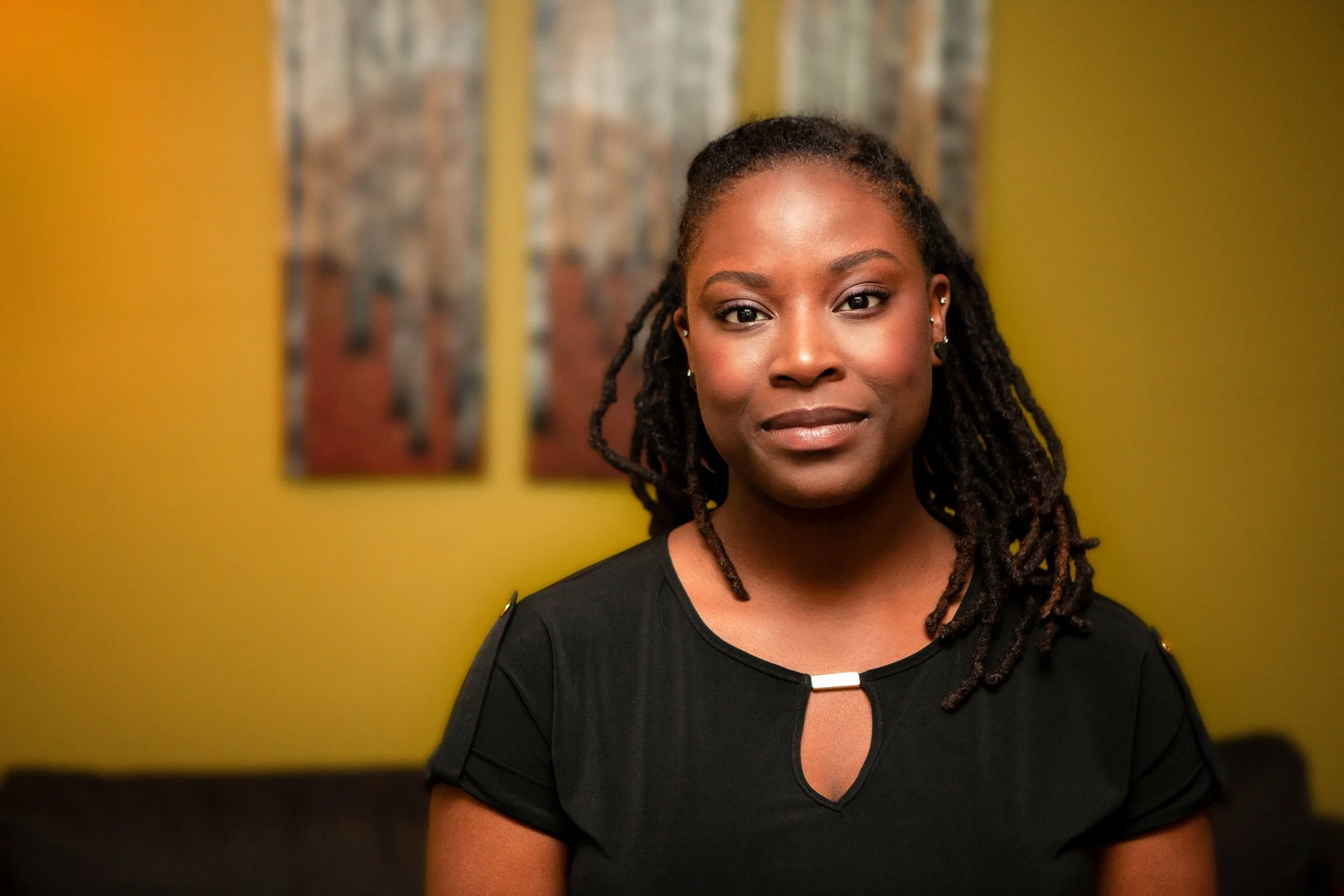 A woman with dark skin and dreadlocks wearing a black top, standing in front of a yellow wall with two paintings in the background.