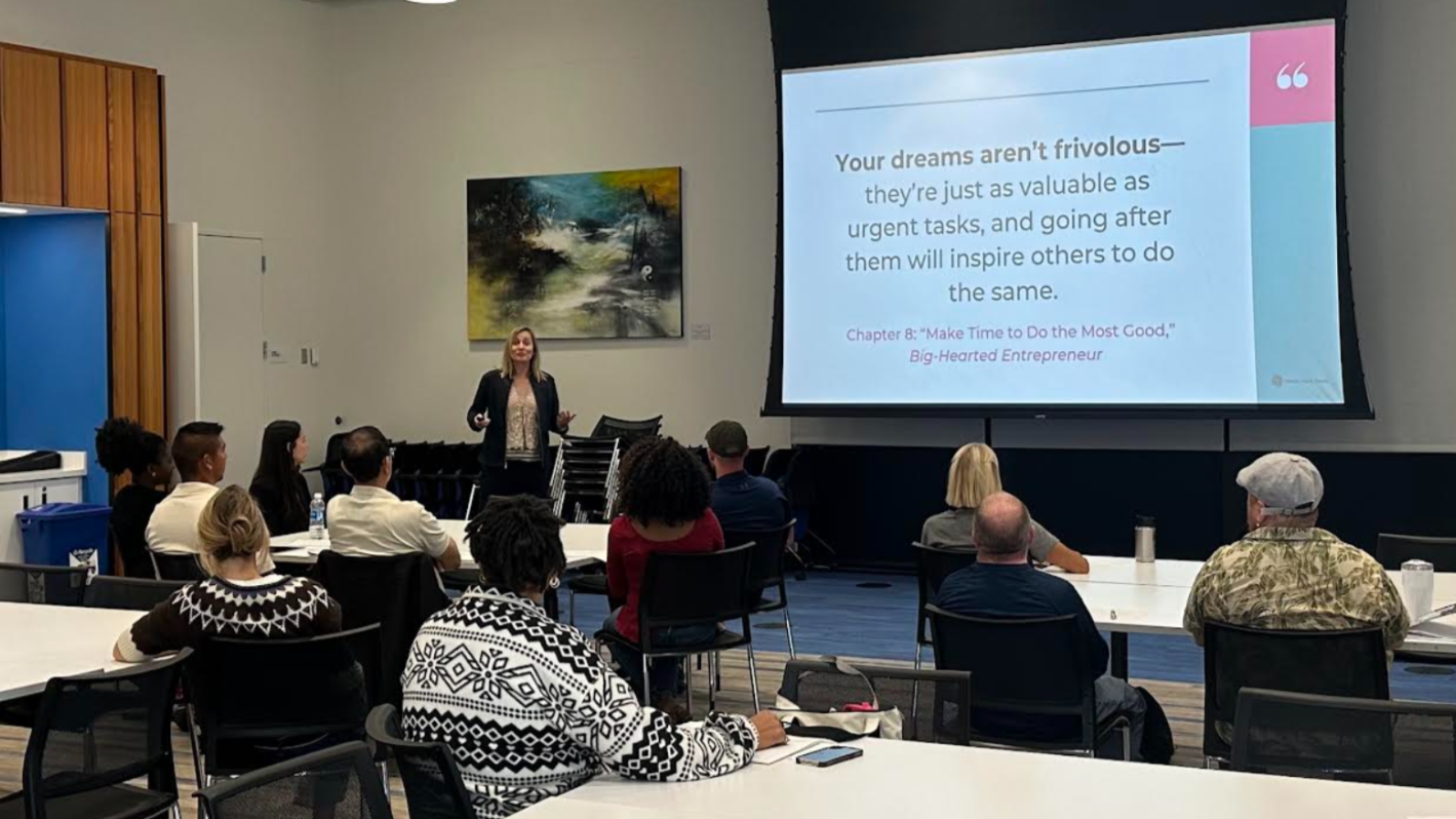 A woman is giving a presentation in a conference room with a large screen displaying a motivational quote, and an audience seated at tables listening.