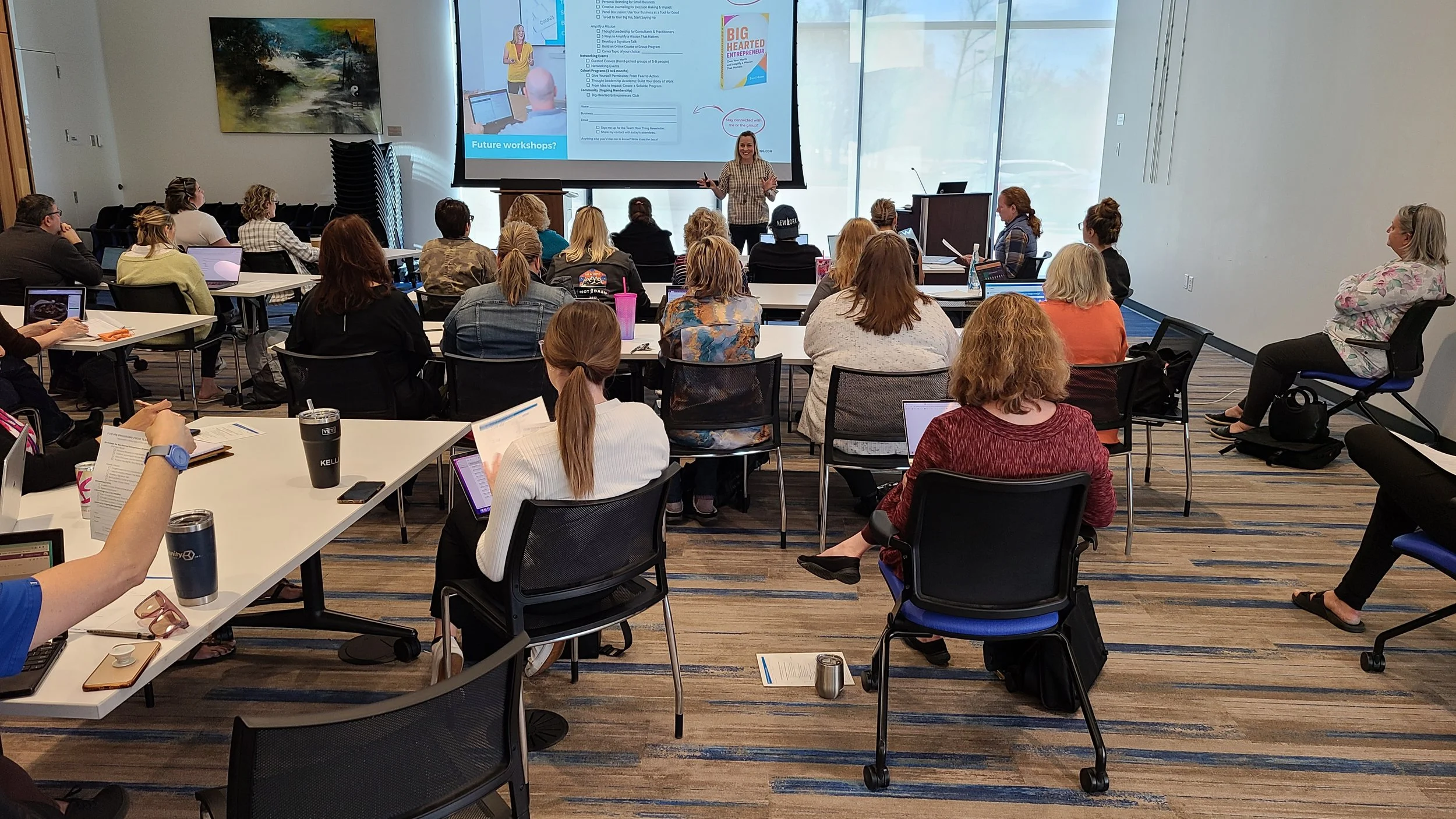A woman is giving a presentation in a conference room to a group of adults seated at tables with laptops and beverages. A large screen behind her displays slides and a colorful book cover, and a painting hangs on the wall to the left.