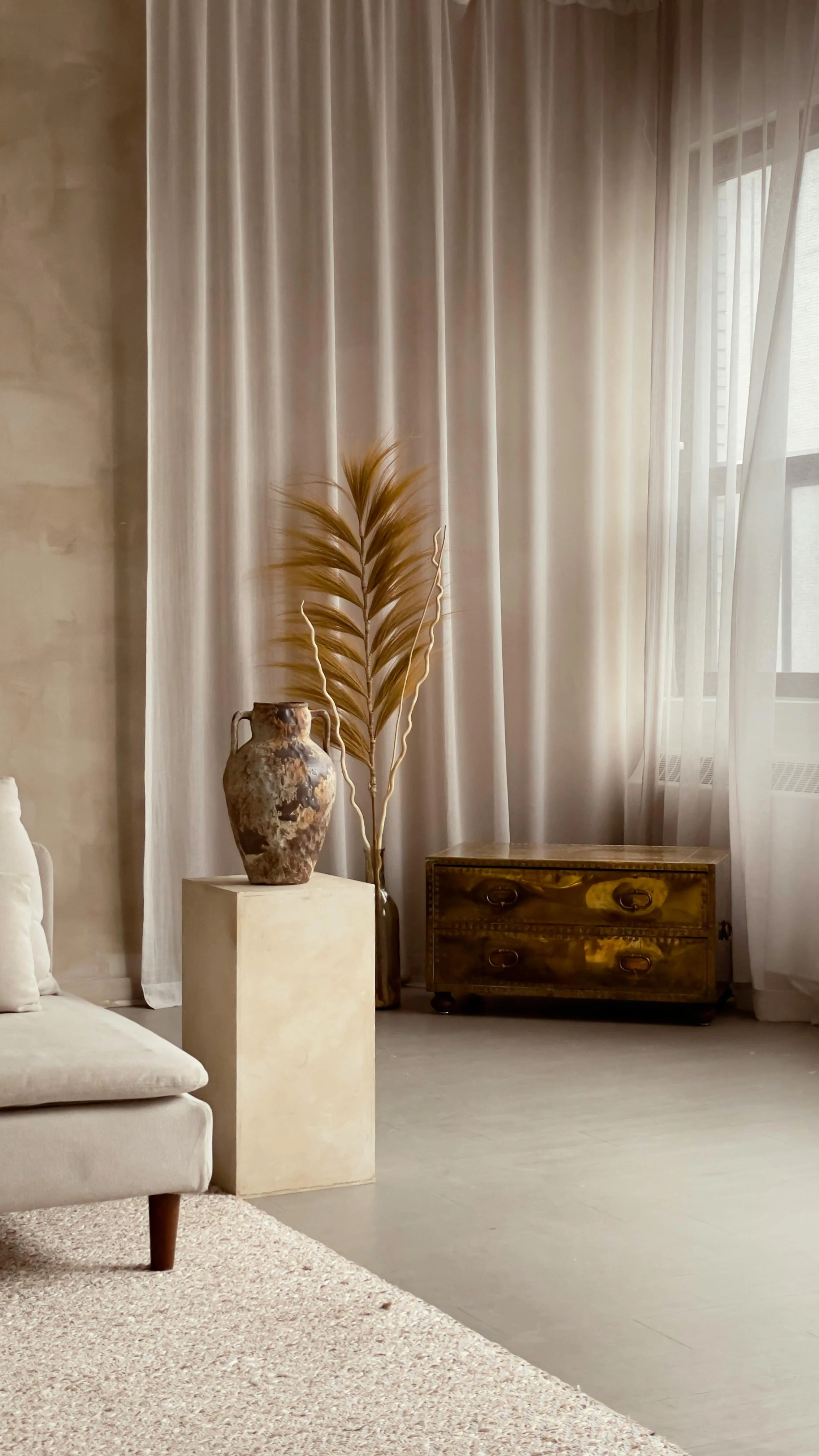 Living room corner with beige sofa, textured beige rug, pottery vase, dried palm leaf, small beige pedestal, wooden side table, sheer curtains, and window with city view. Showing the interior part of Euki Studio