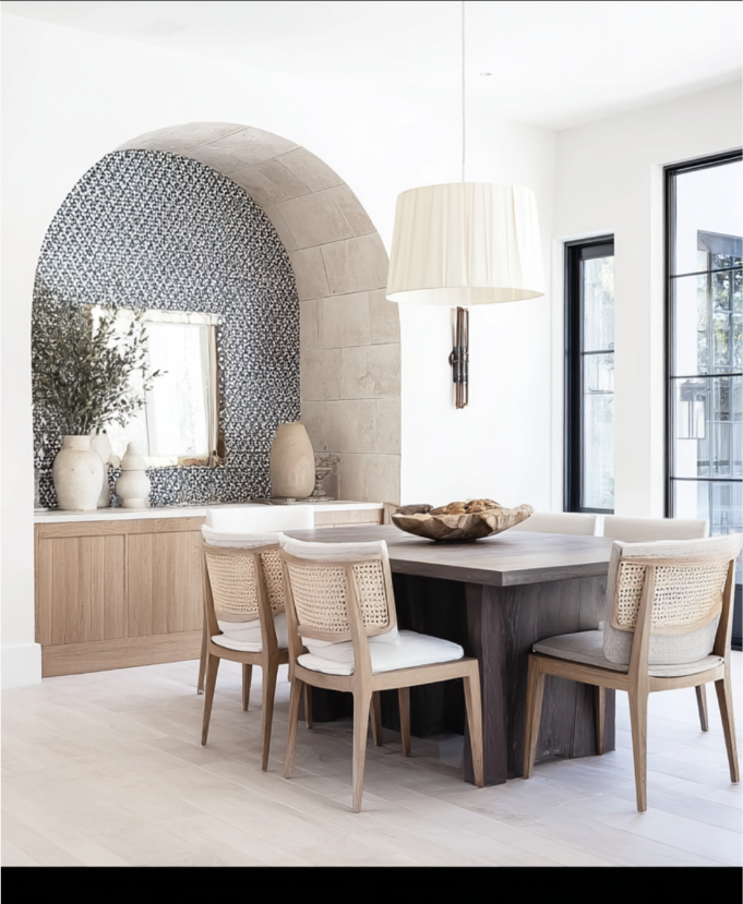 Modern dining room with a dark wooden table, six chairs with wicker backs, a large cream pendant light, and decorative elements like vases and a mirror, with a patterned archway and windows letting in natural light.