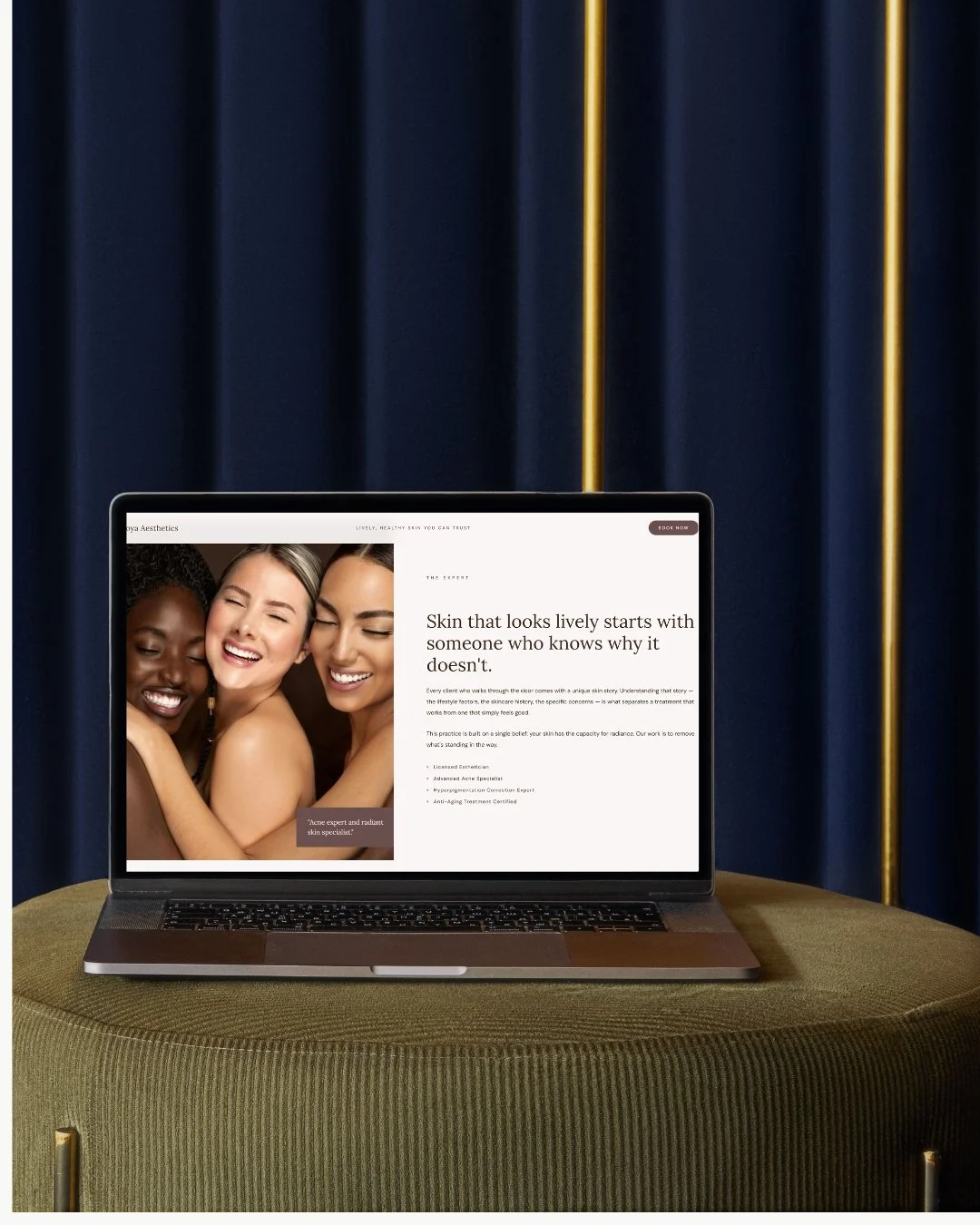 A laptop on a green textured table displays a website with a photo of three smiling women and text about skincare and skin professionals, set against a dark blue curtain background with gold accents.