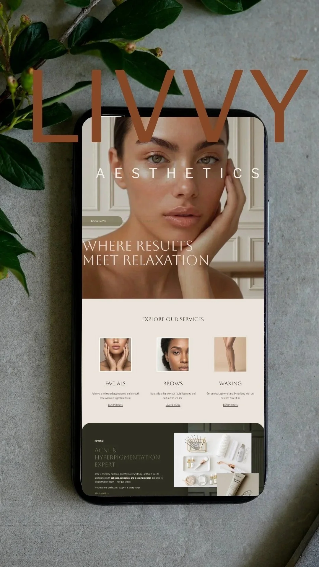 A smartphone displaying a website for LIVVY Aesthetics, featuring a close-up of a woman's face, and service options for facials, brows, and waxing, placed on a gray surface with green leaves nearby.