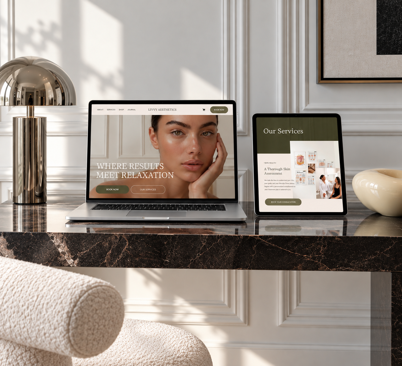A marble desk with a laptop and tablet displaying a skincare website, a chrome desk lamp, a white vase, and part of a beige armchair in a bright room with white paneled walls and framed art.