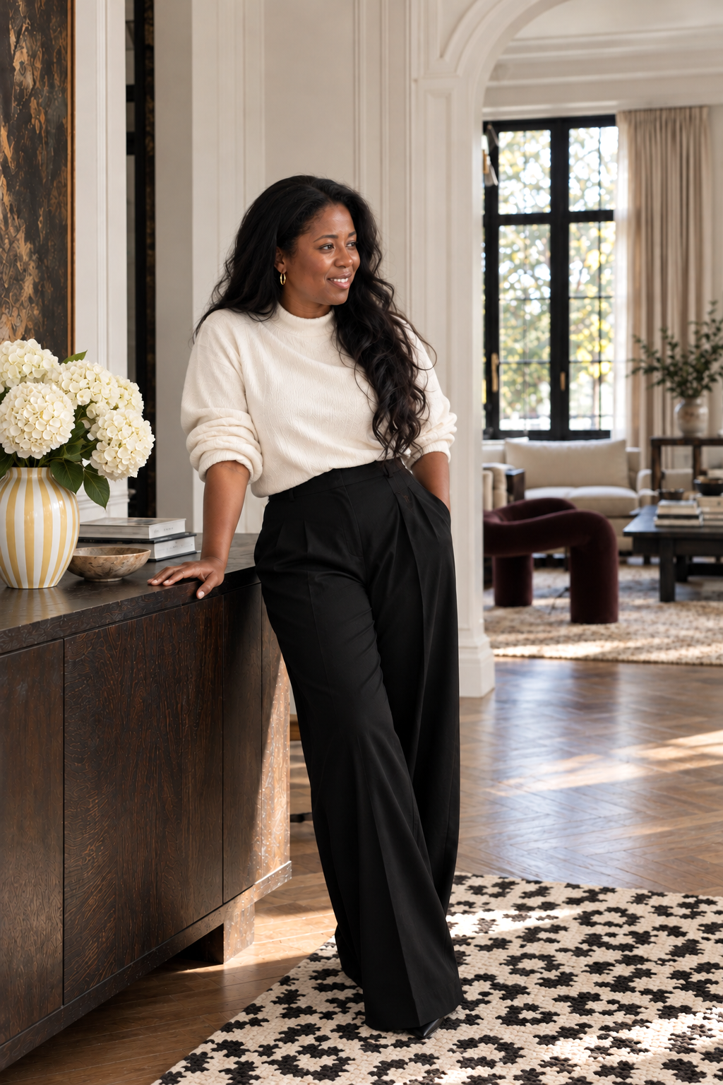 A woman with long dark hair, wearing a white sweater and black wide-leg pants, is leaning on a wooden sideboard with white flowers in a yellow striped vase inside a spacious, well-lit living room.