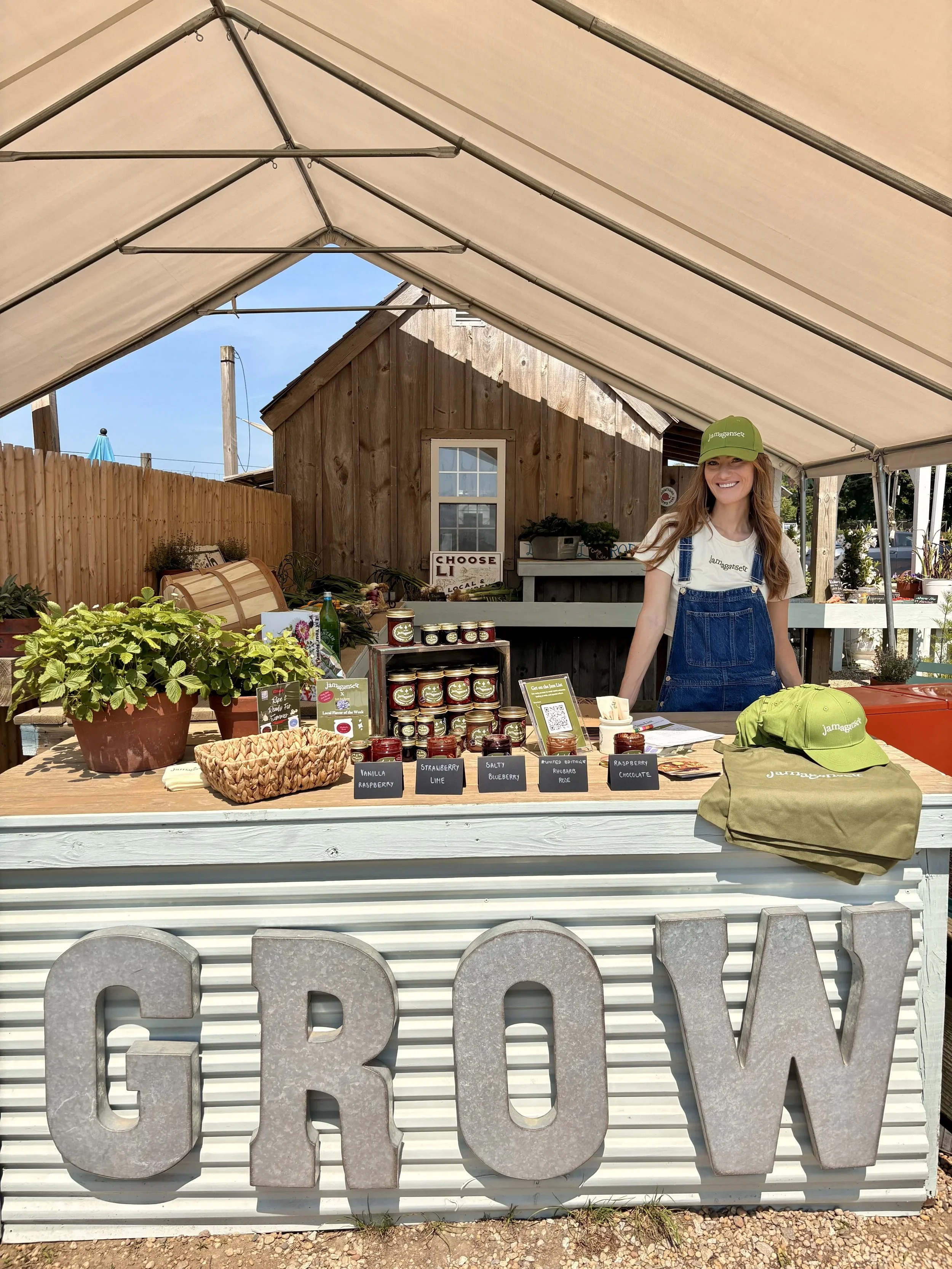 A woman smiling at a farmers market stand selling jams and preserves, with a wooden shed, plants, and a large metal 'GROW' sign in the background. She is wearing a green cap and denim apron. The stand has jars of jam labeled with flavors like vanilla raspberry, strawberry lime, blueberry, raspberry chocolate, and raspberry rhubarb rose.