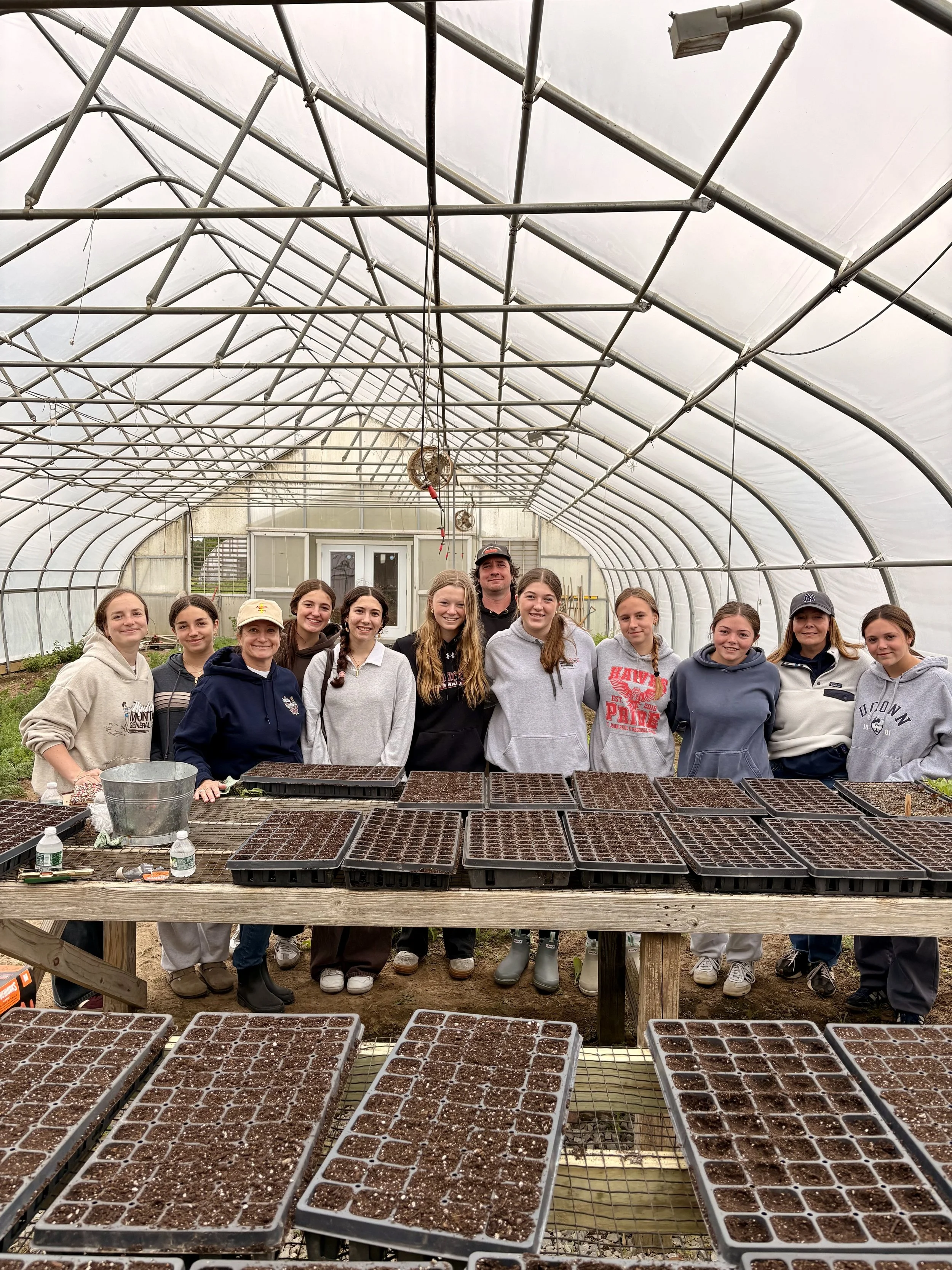 Group of young people and a man standing inside a greenhouse with seed trays on tables in front of them.